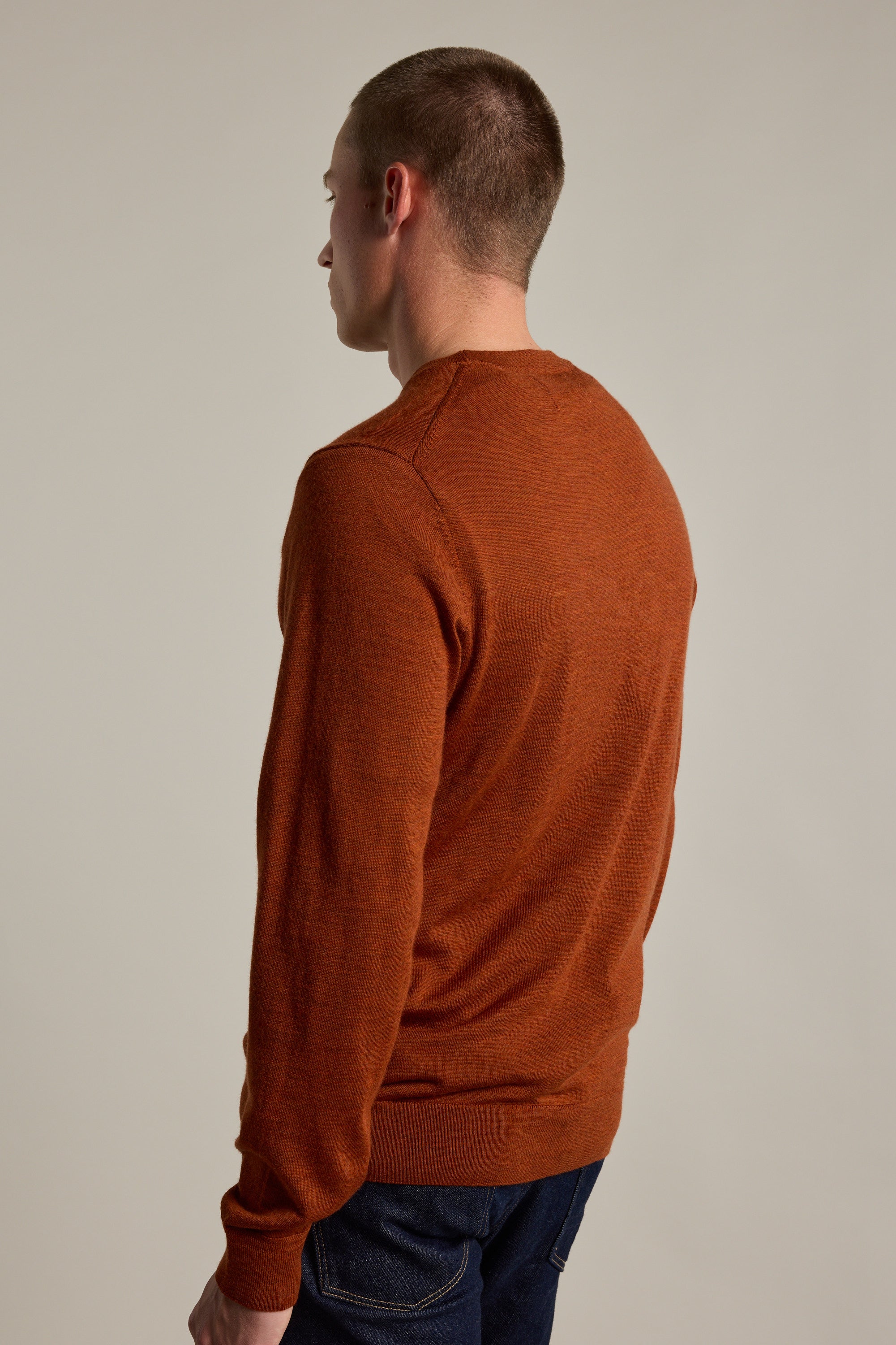 A person with short hair, wearing a rust Skyland ZQrx Merino Crew by Barkers and dark jeans, stands facing away from the camera against a neutral background.