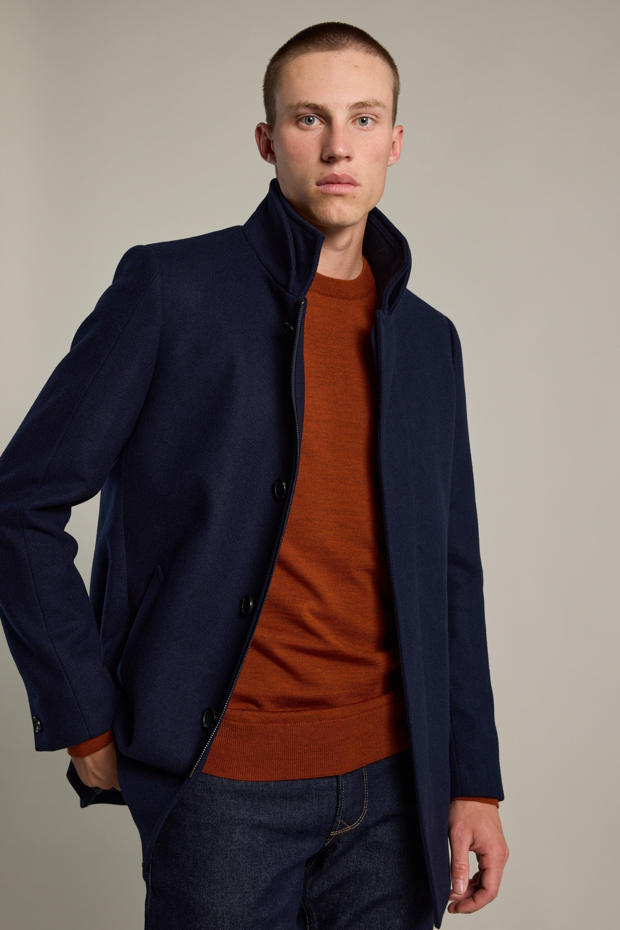 A young man with short hair wears a Barkers Skyland ZQrx Merino Crew in burnt orange under a navy coat and dark jeans, standing against a plain light background with one hand in his pocket.