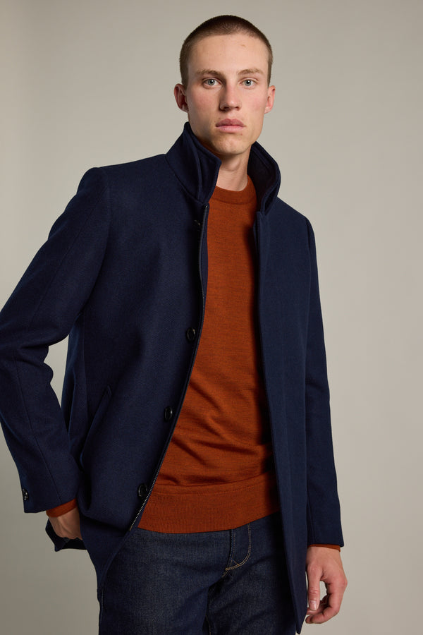 A young man with short hair wears a Barkers Skyland ZQrx Merino Crew in burnt orange under a navy coat and dark jeans, standing against a plain light background with one hand in his pocket.