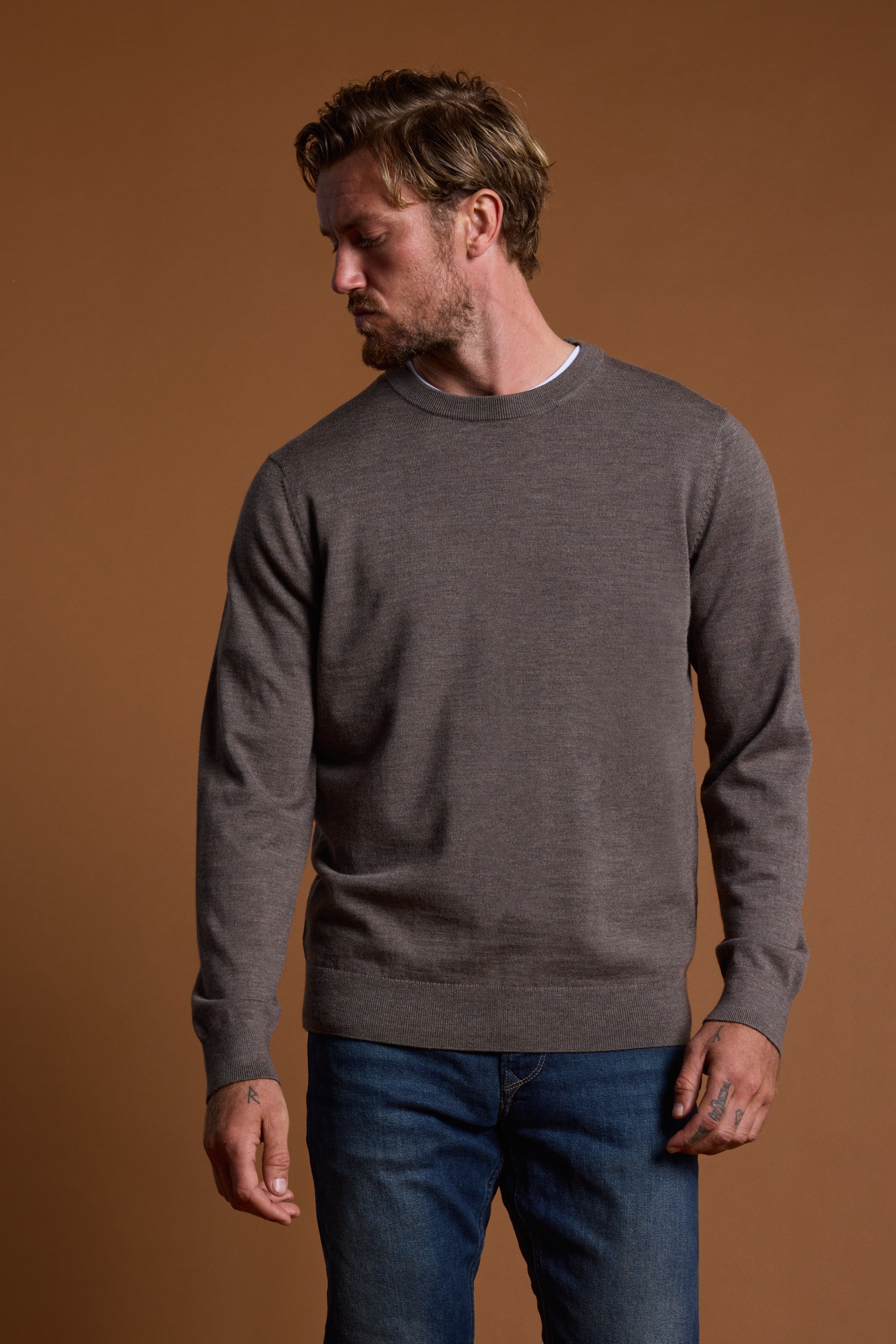A man with light brown hair and a short beard wears the Barkers Skyland ZQrx Merino Crew sweater and blue jeans, standing against a plain brown background, looking down to his left.