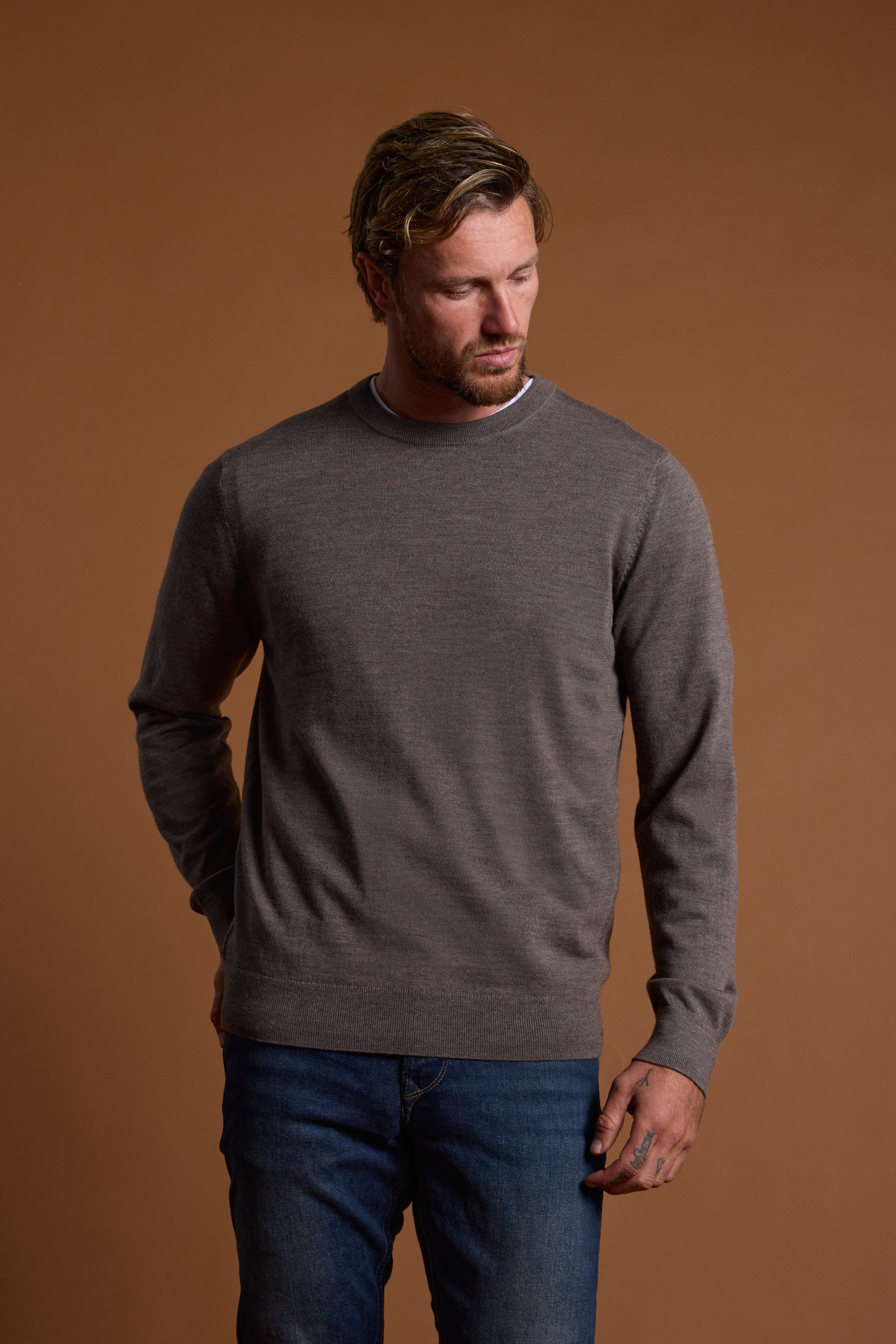 A man with light brown hair and a short beard wears the Barkers Skyland ZQrx Merino Crew in gray and blue jeans. He stands against a plain brown background, looking down to his left with one hand in his pocket.