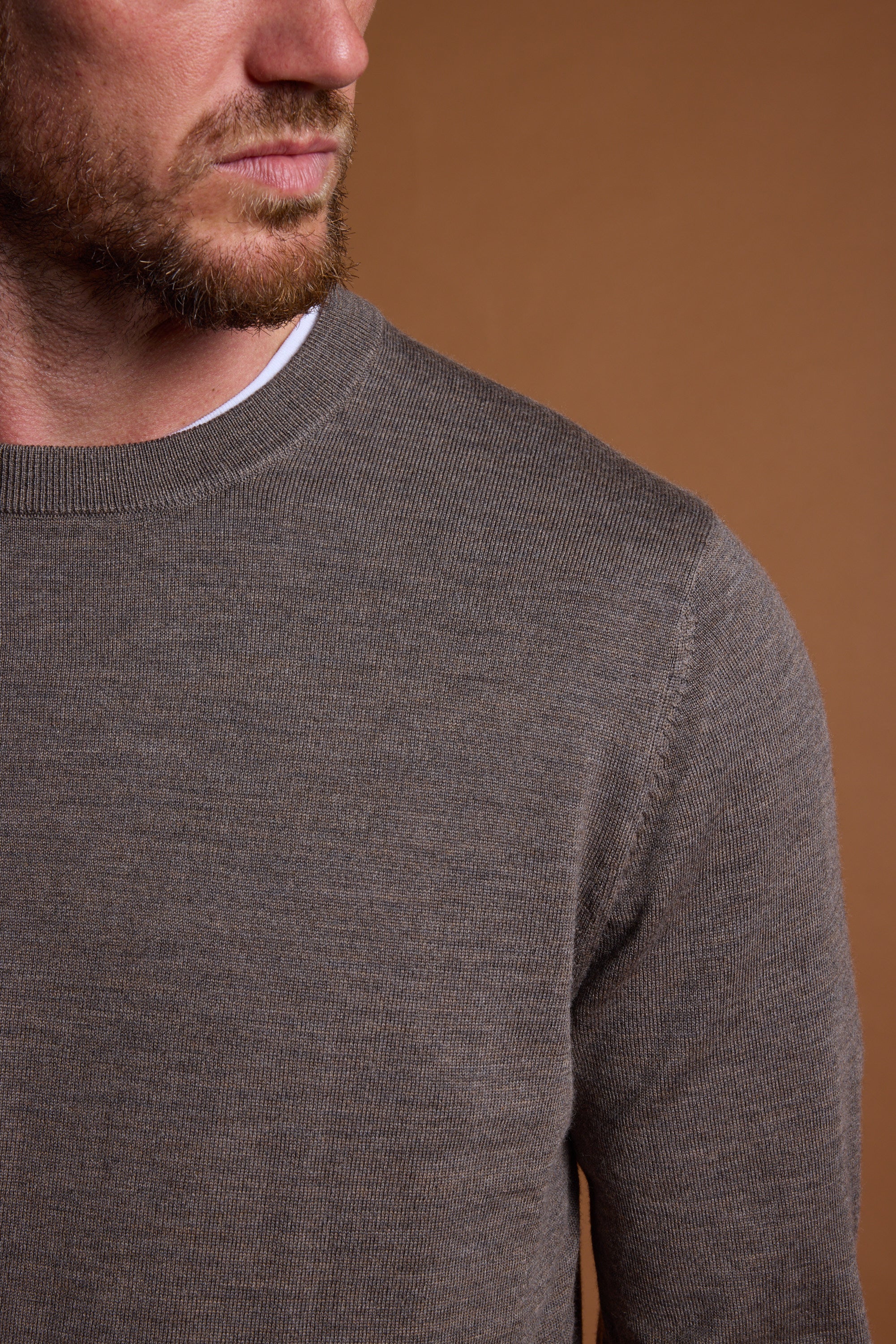 A man with a short beard and mustache wears the Barkers Skyland ZQrx Merino Crew, a brown merino wool sweater, over a white shirt. Shown from the shoulders up against a plain brown background, his face is partially visible, looking left.