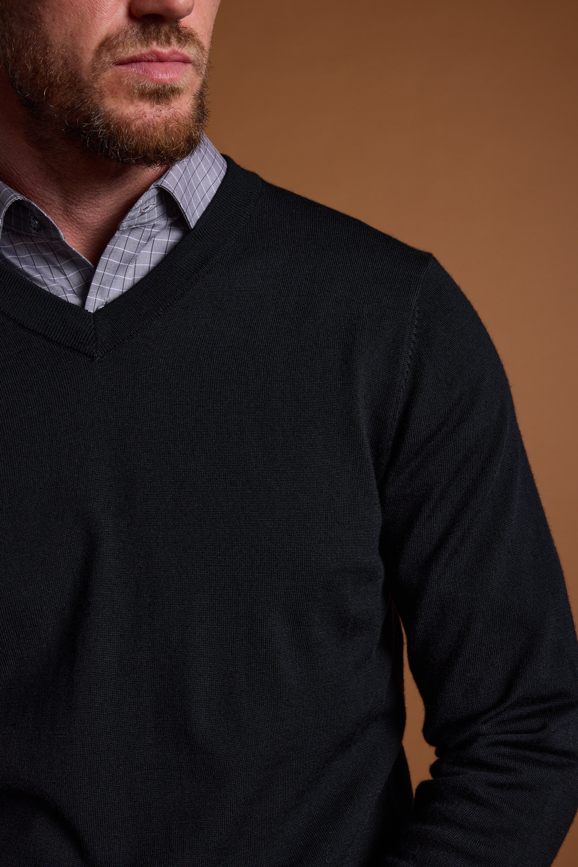 A man in the Barkers Skyland ZQrx Merino Vee, a dark V-neck sweater made from traceable merino, layered over a checkered collared shirt, is shown from the shoulders up with his face partly visible against a brown background.