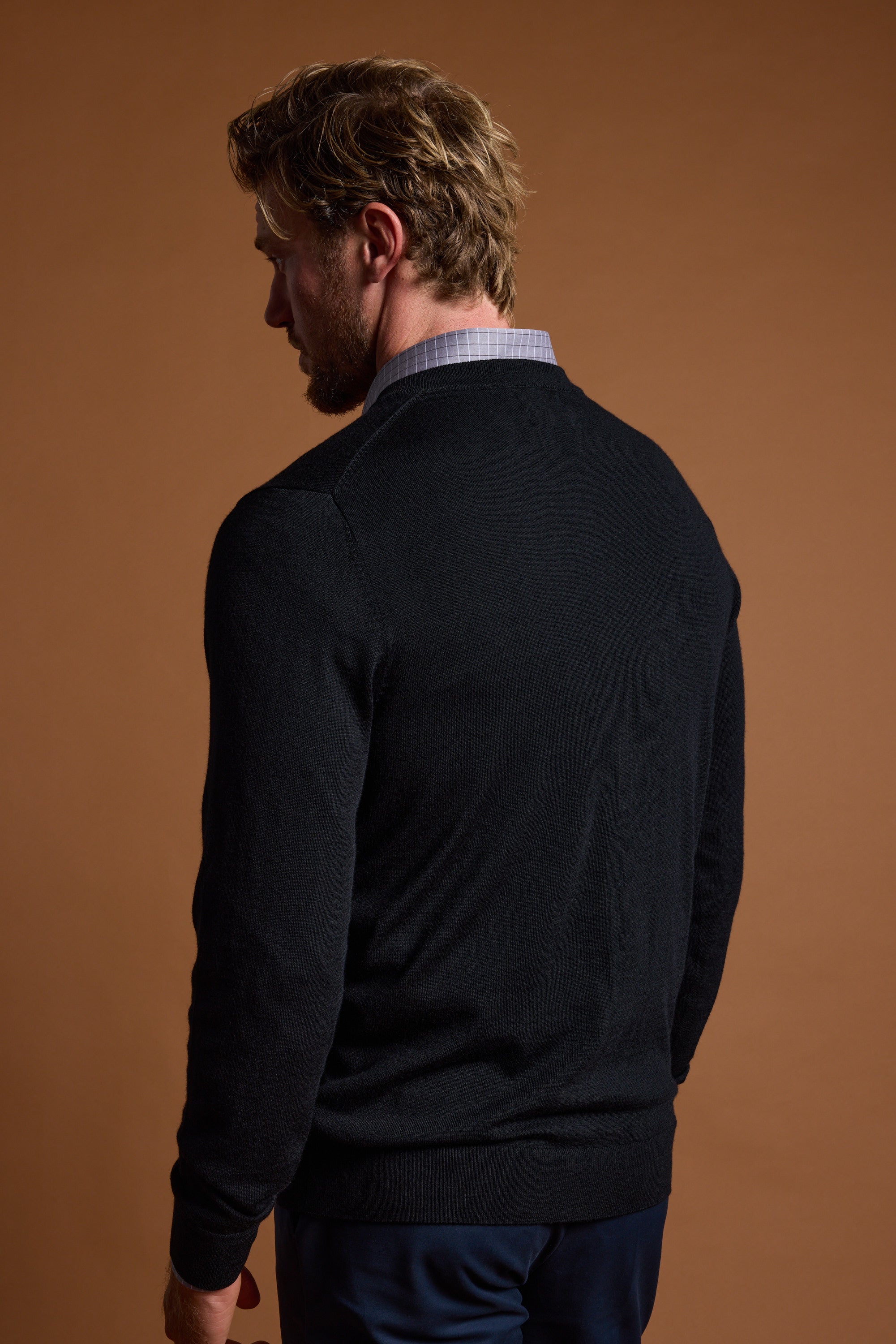 A man with light brown hair wears a Barkers Skyland ZQrx Merino Vee, a black New Zealand merino wool sweater over a collared shirt, standing with his back to a plain brown background.