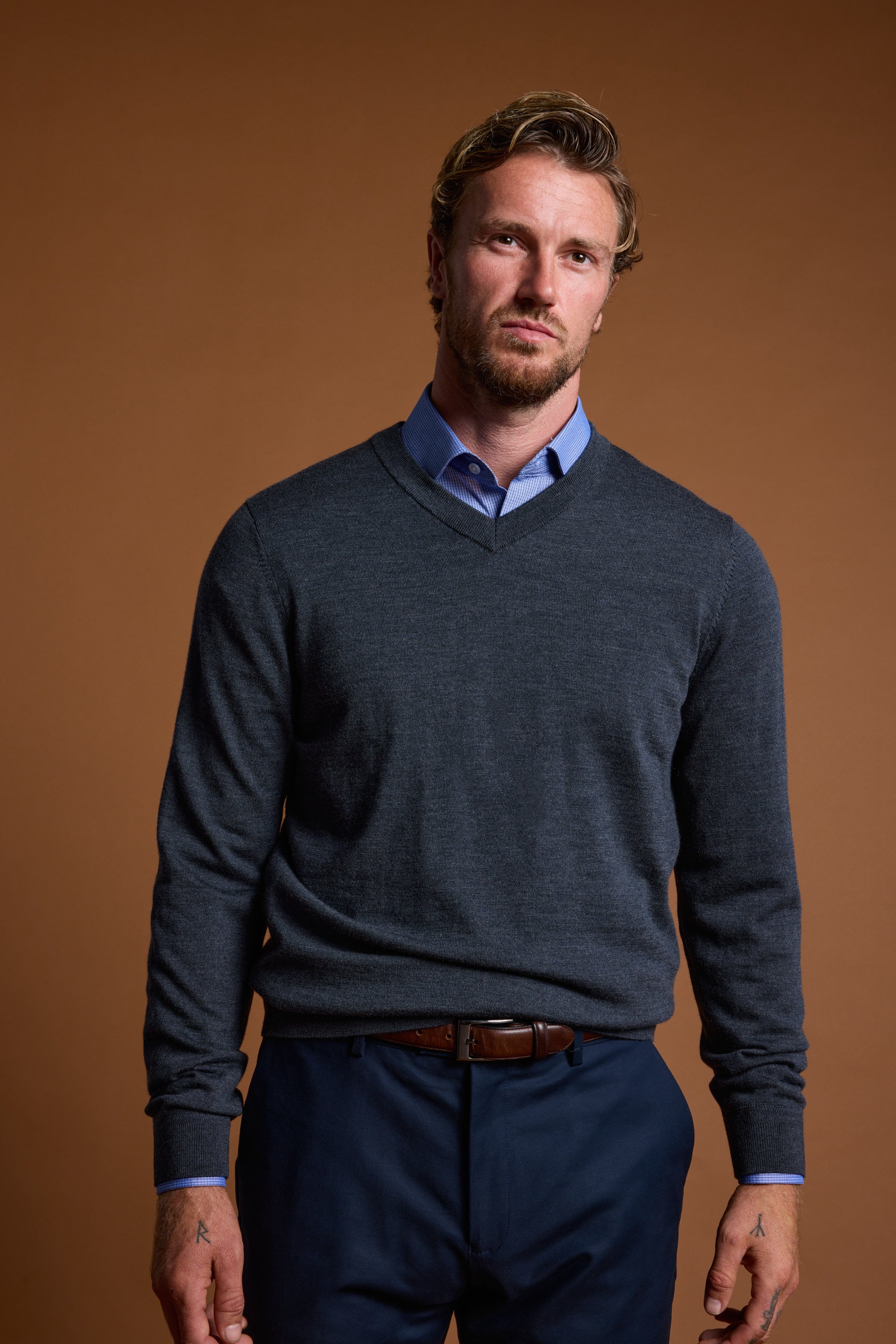 A man with short, wavy blond hair and a beard stands against a brown background, wearing Barkers' Skyland ZQrx Merino Vee sweater over a blue shirt and dark pants, looking seriously at the camera.