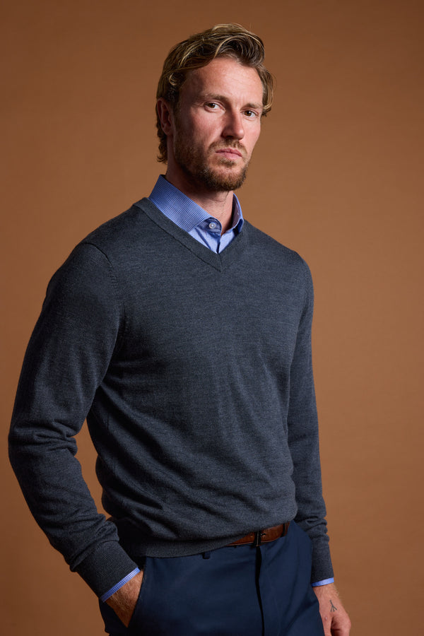 A man with light brown hair and a short beard wears a blue dress shirt and the Barkers Skyland ZQrx Merino Vee sweater, standing with one hand in his pocket against a plain brown background.