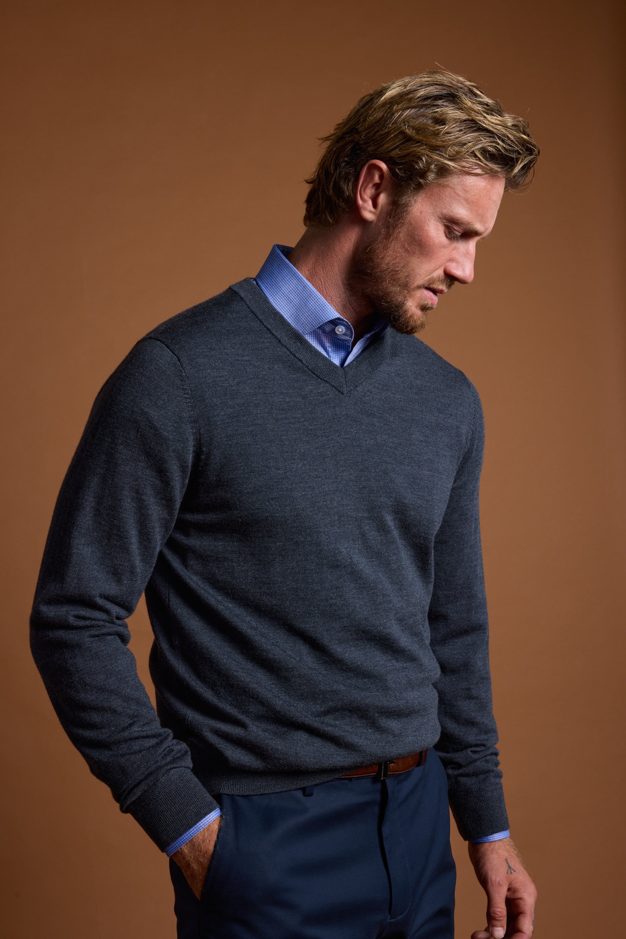 A man with blond hair and a short beard models the Barkers Skyland ZQrx Merino Vee, a dark gray, breathable sweater made from ethically sourced New Zealand merino wool, layered over a blue shirt. He stands against a brown background looking down.
