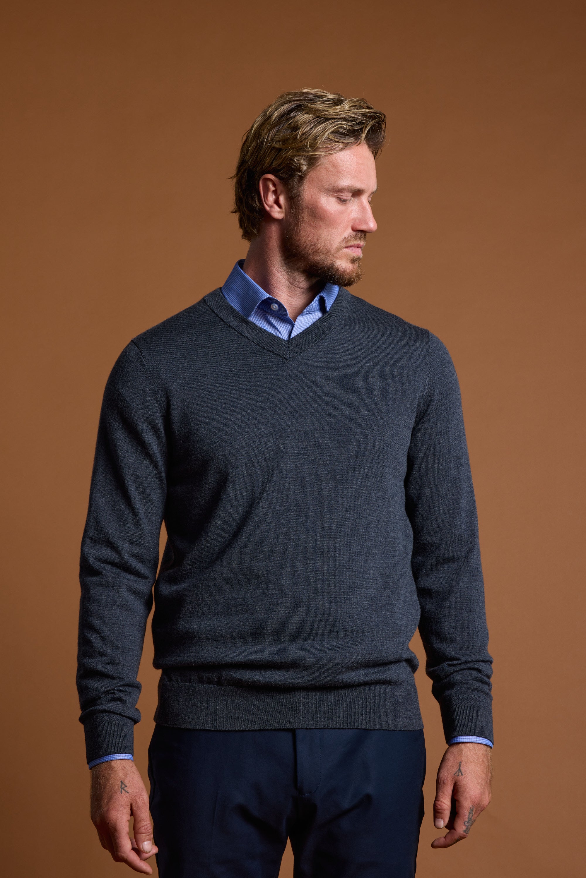 A blond man with a beard wears the Barkers Skyland ZQrx Merino Vee, a blue-collared shirt under a dark gray V-neck sweater made from breathable New Zealand merino wool, standing against a plain brown background and looking left.