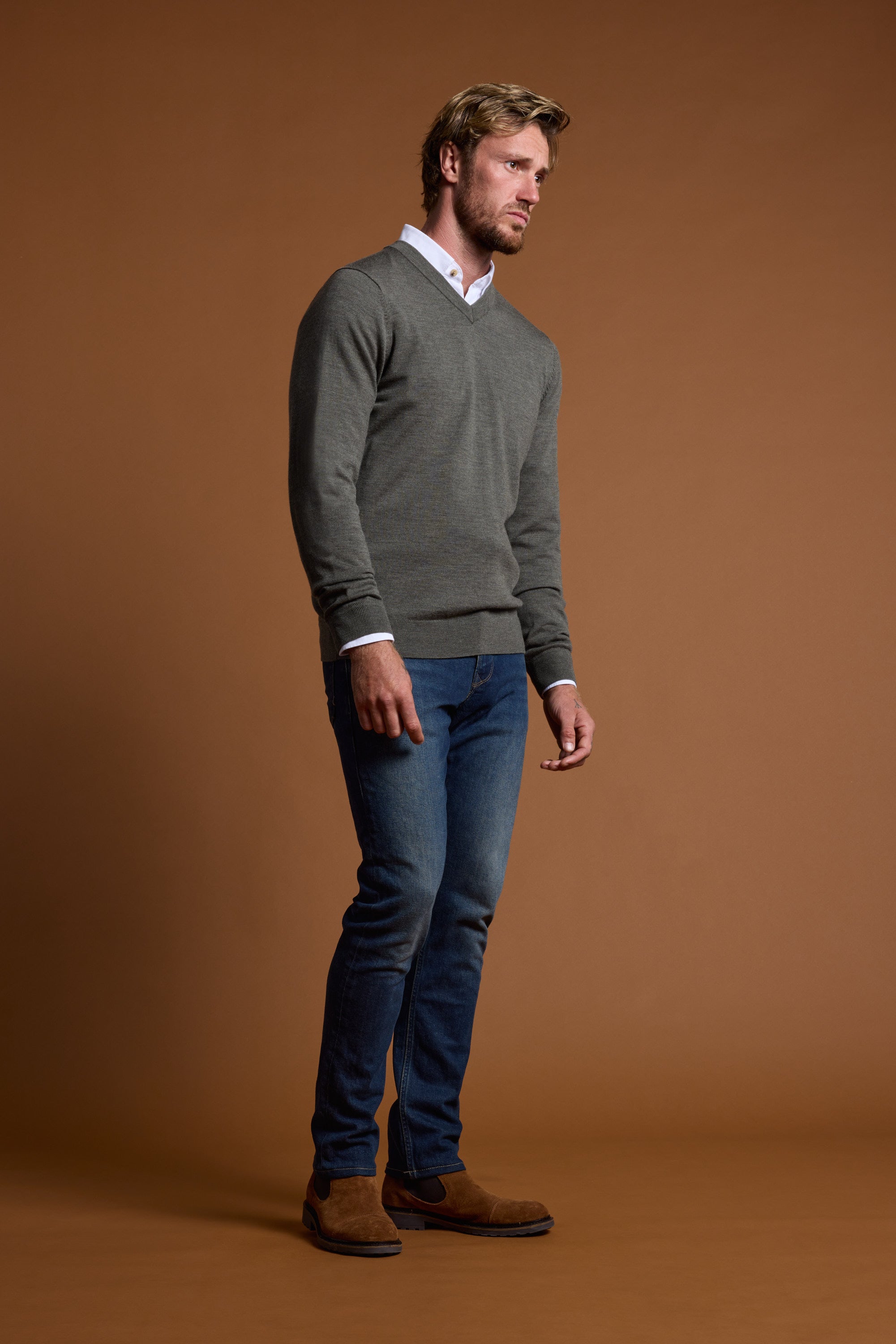 A man with light hair and a beard, looking to the side, wears the Barkers Skyland ZQrx Merino Vee sweater over a white shirt, blue jeans, and brown suede shoes against a plain brown background.