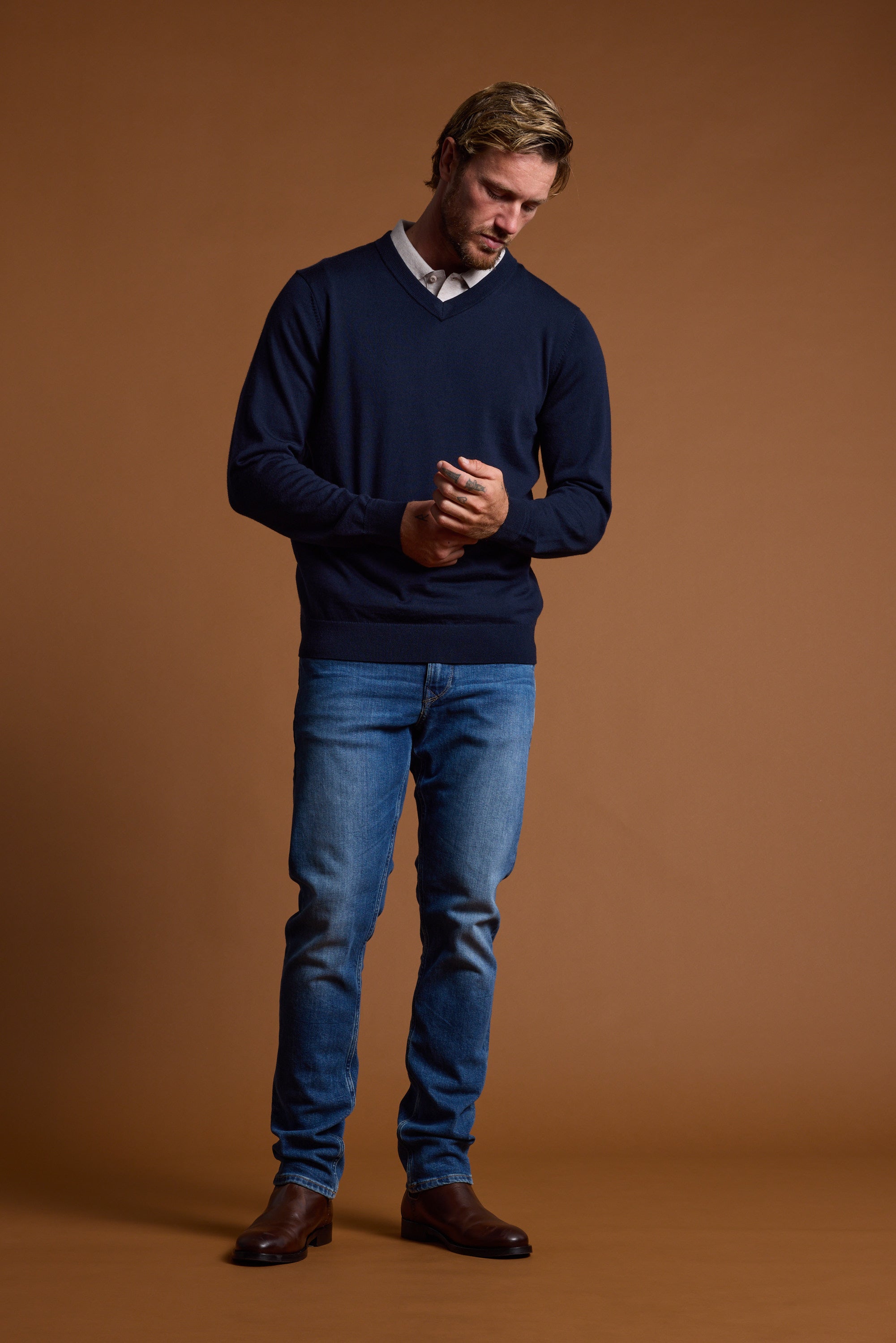 A man stands against a brown background, wearing the Barkers Skyland ZQrx Merino Vee sweater over a white collared shirt, blue jeans, and brown boots. He looks down with his hands clasped in front of him.