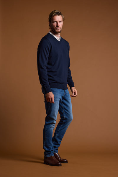 A blonde-bearded man stands against a brown background, wearing the Barkers Skyland ZQrx Merino Vee sweater over a light collared shirt, blue jeans, and brown shoes, looking at the camera with a neutral expression.
