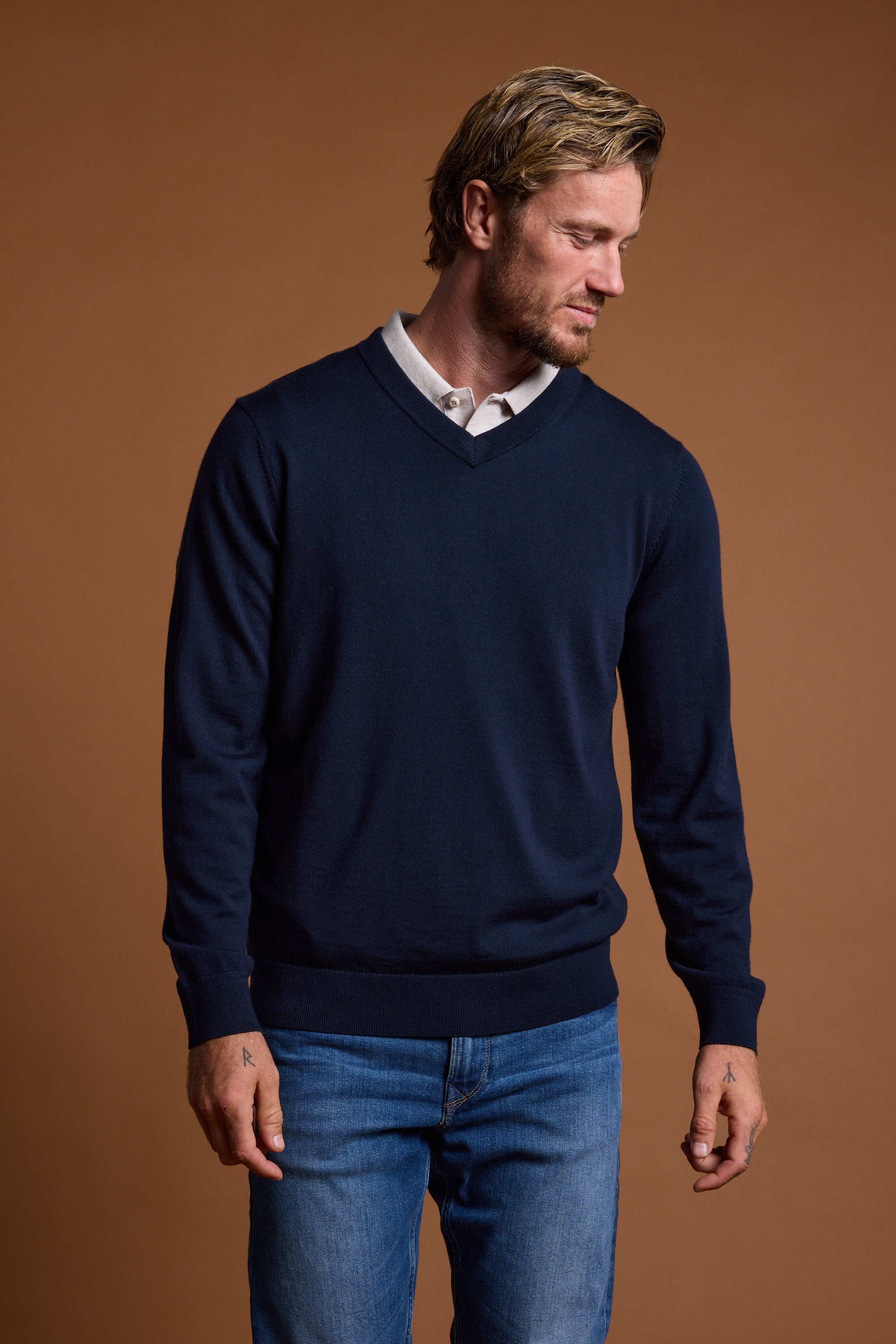 A man with short blond hair and a beard stands against a brown background, wearing the Barkers Skyland ZQrx Merino Vee—a navy blue New Zealand merino wool V-neck sweater—over a white shirt and blue jeans, looking down to his left.