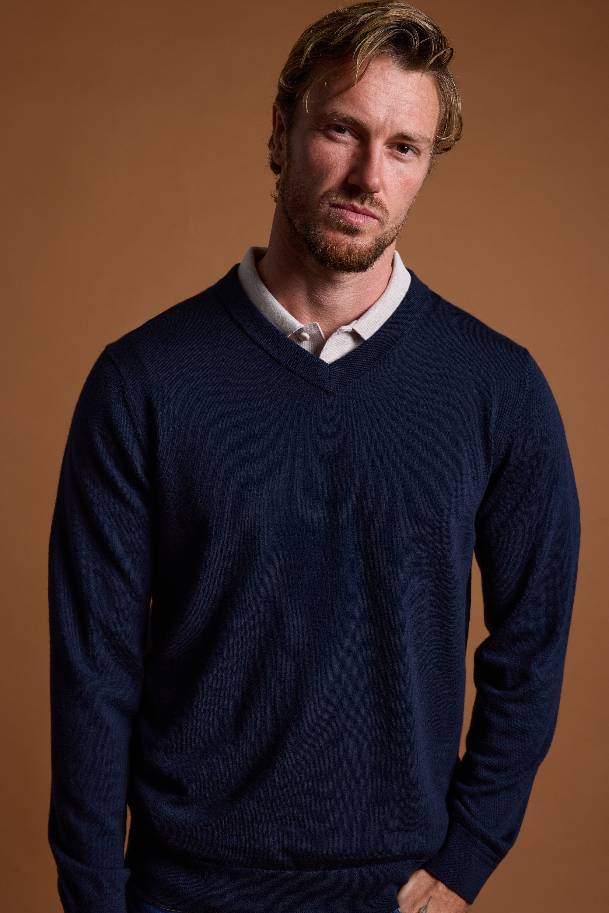 A man with light brown hair and a short beard stands against a brown background, wearing the Barkers Skyland ZQrx Merino Vee— a navy V-neck sweater made from New Zealand merino wool—over a light collared shirt. He looks neutrally at the camera.