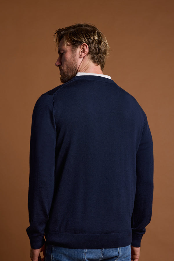 A man with light brown hair, wearing Barkers' Skyland ZQrx Merino Vee navy blue sweater and blue jeans, stands facing away from the camera against a brown background.