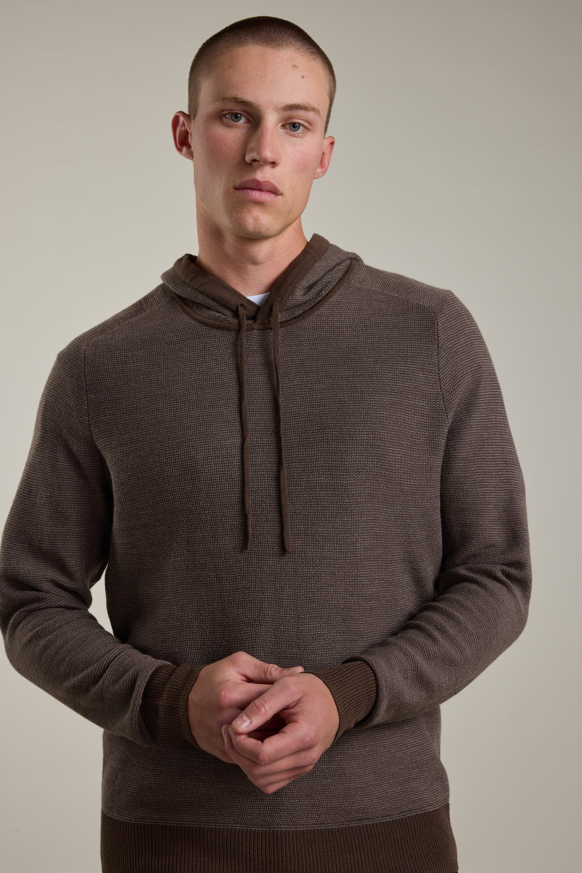 A person stands against a plain background wearing the Barkers Roxbury Merino Hoody in brown, hands loosely clasped at their waist and a neutral expression, highlighting this lightweight sweater’s effortless style.