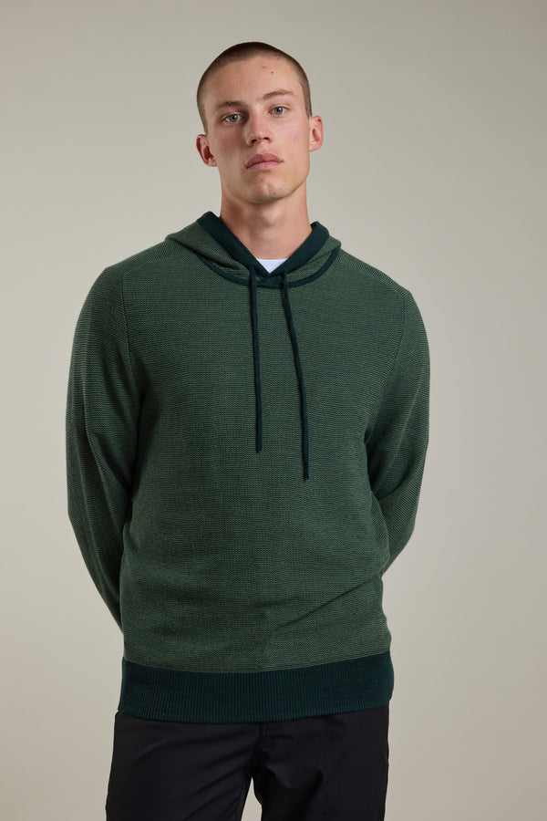 A young man with short hair stands against a plain light background, calmly looking at the camera with his hands behind his back, wearing Barkers’ Roxbury Merino Hoody and dark pants.