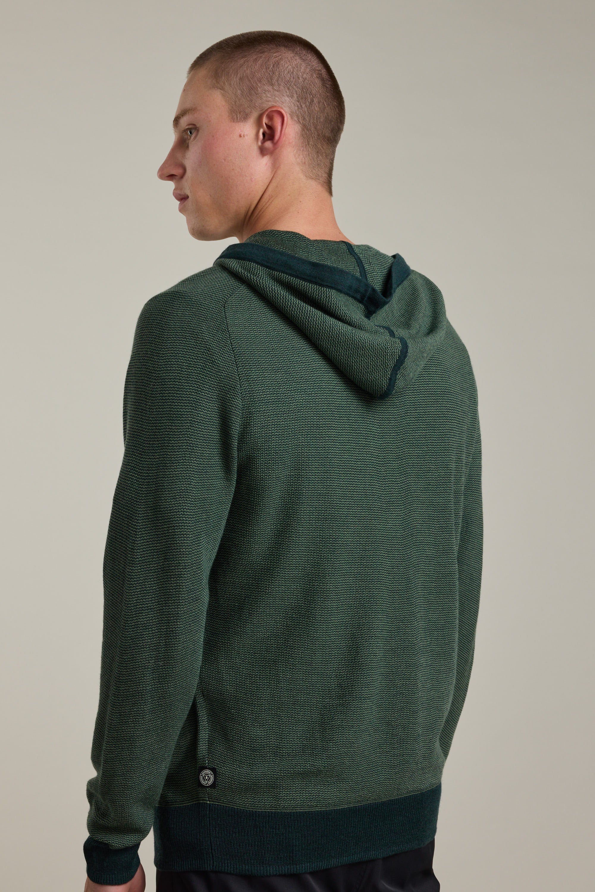 A person with short hair wears a dark green Barkers Roxbury Merino Hoody, standing against a plain light background and looking over their left shoulder, showing the back and side profile of the lightweight knit sweater.