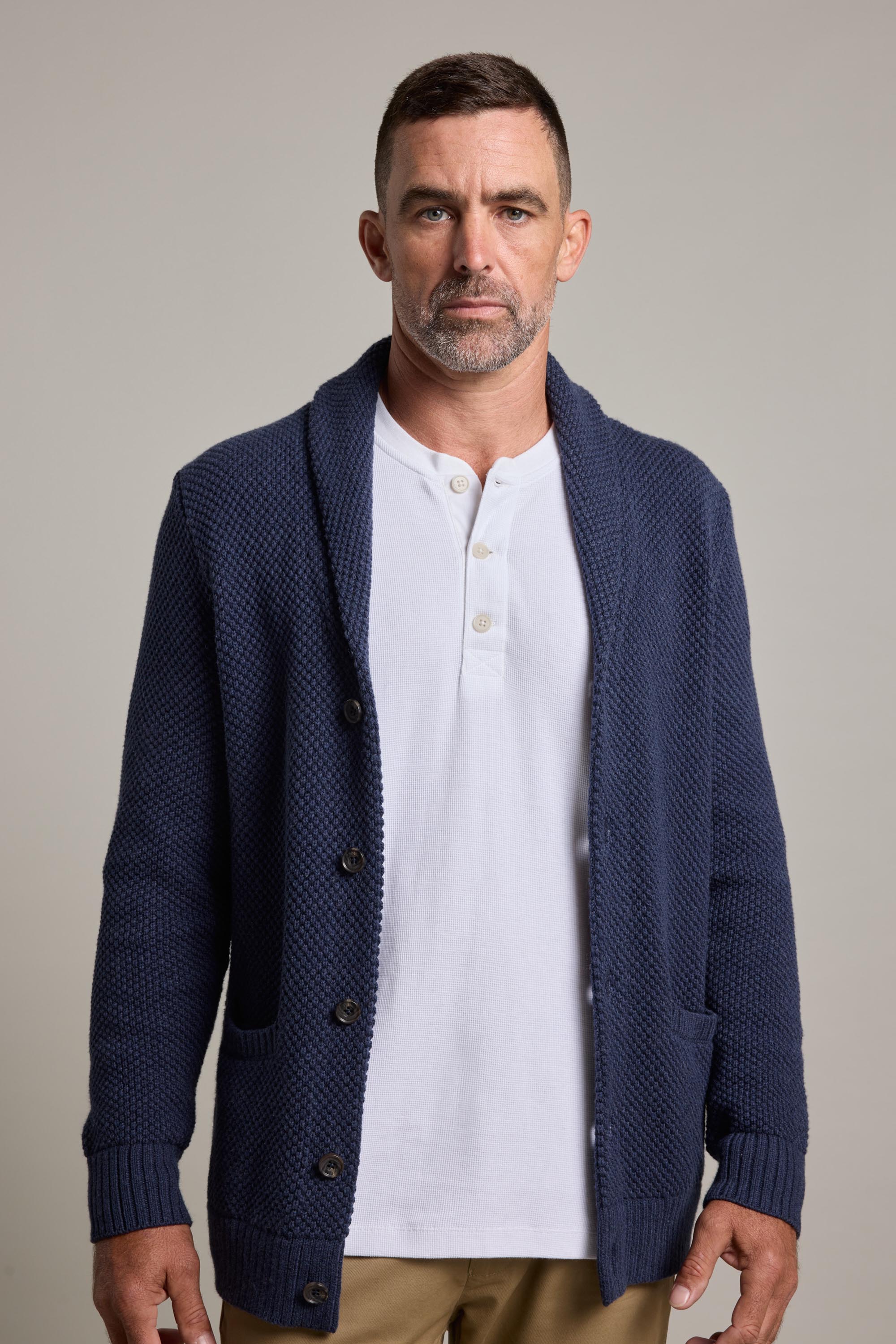 A man with short dark hair and a trimmed beard models Barkers’ Brighton Cotton Shawl Cardi in navy over a white henley and tan pants, set against a plain light gray background—perfect for winter layering.
