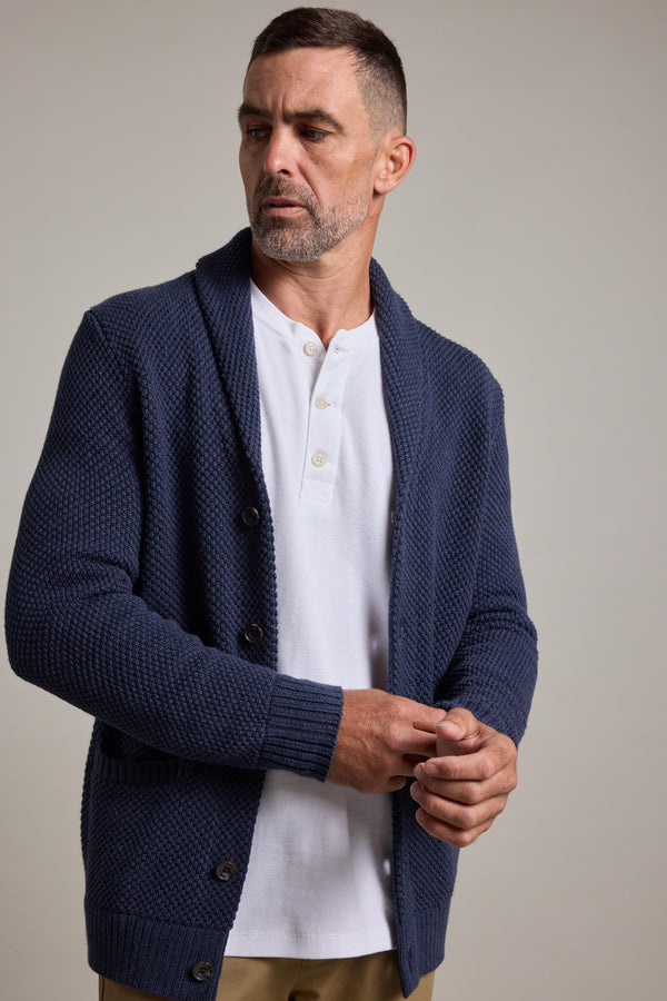 A man with short hair and a beard wears the Barkers Brighton Cotton Shawl Cardi over a white henley shirt and tan pants, standing against a plain light background, looking to his left with hands loosely clasped.