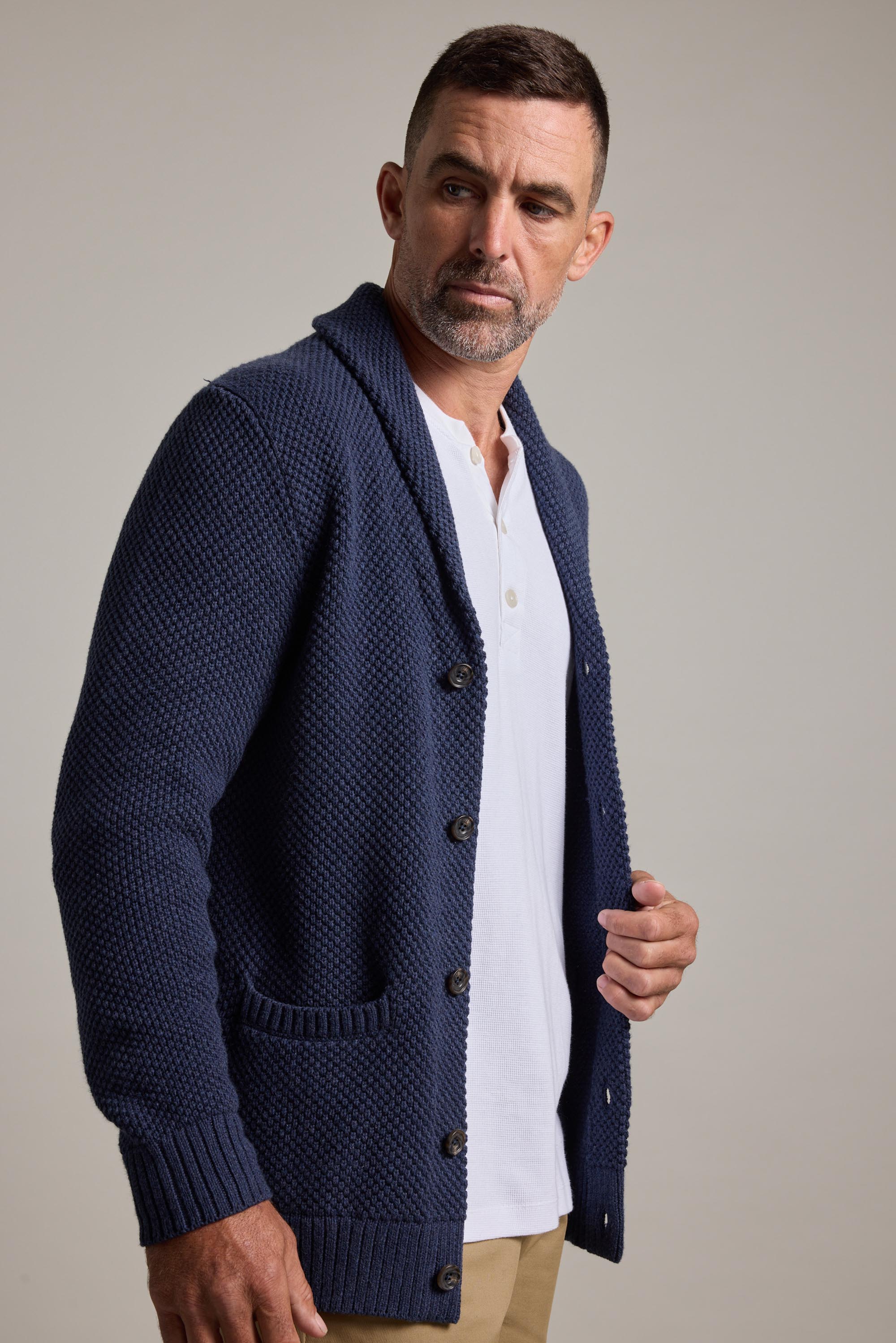 A man with short hair and a trimmed beard wears the Barkers Brighton Cotton Shawl Cardi in textured navy blue over a white shirt and tan pants, standing against a plain light background and looking to his left.