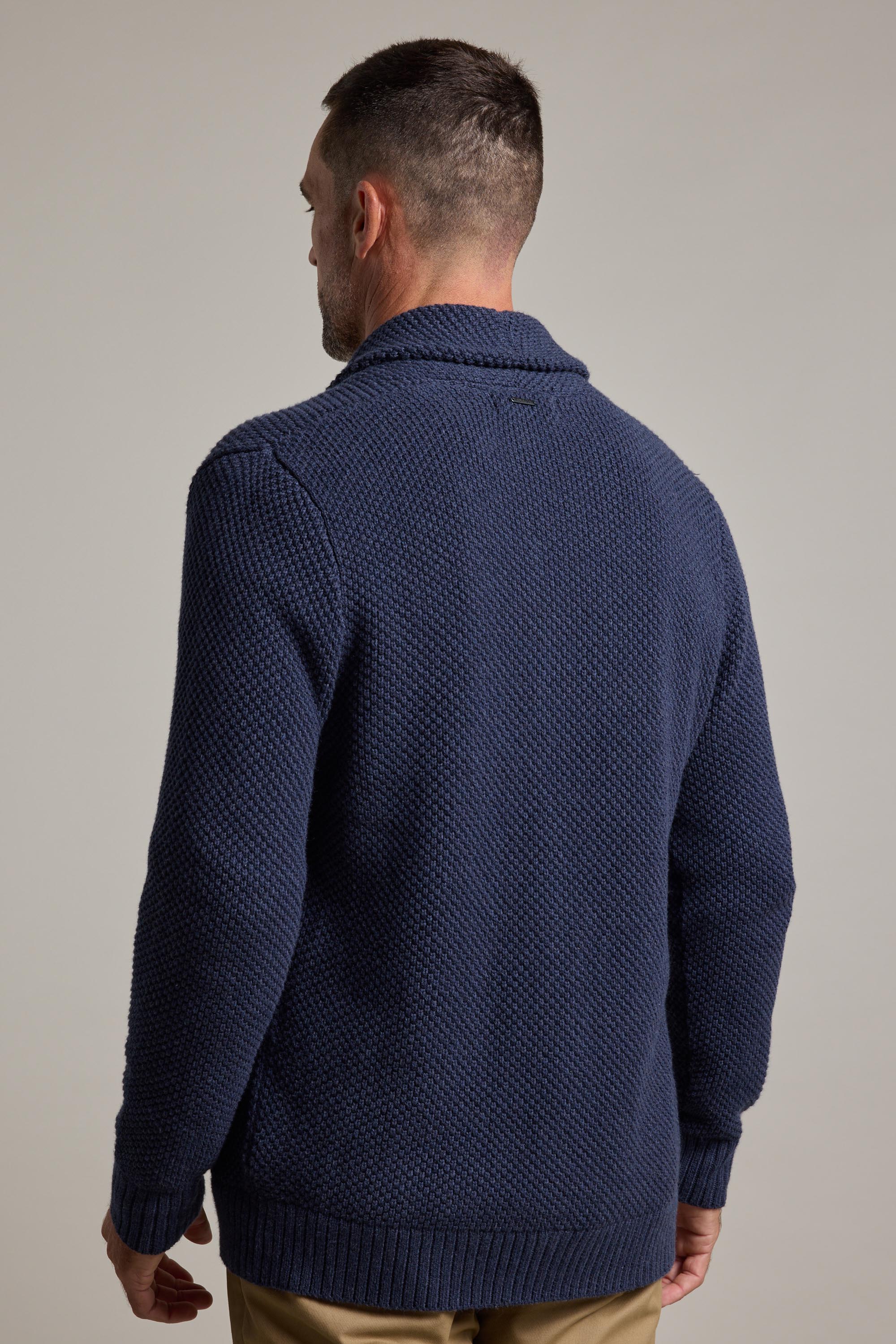 A man with short dark hair is seen from behind, wearing the Barkers Brighton Cotton Shawl Cardi in textured navy blue and tan pants, standing against a plain light gray background.