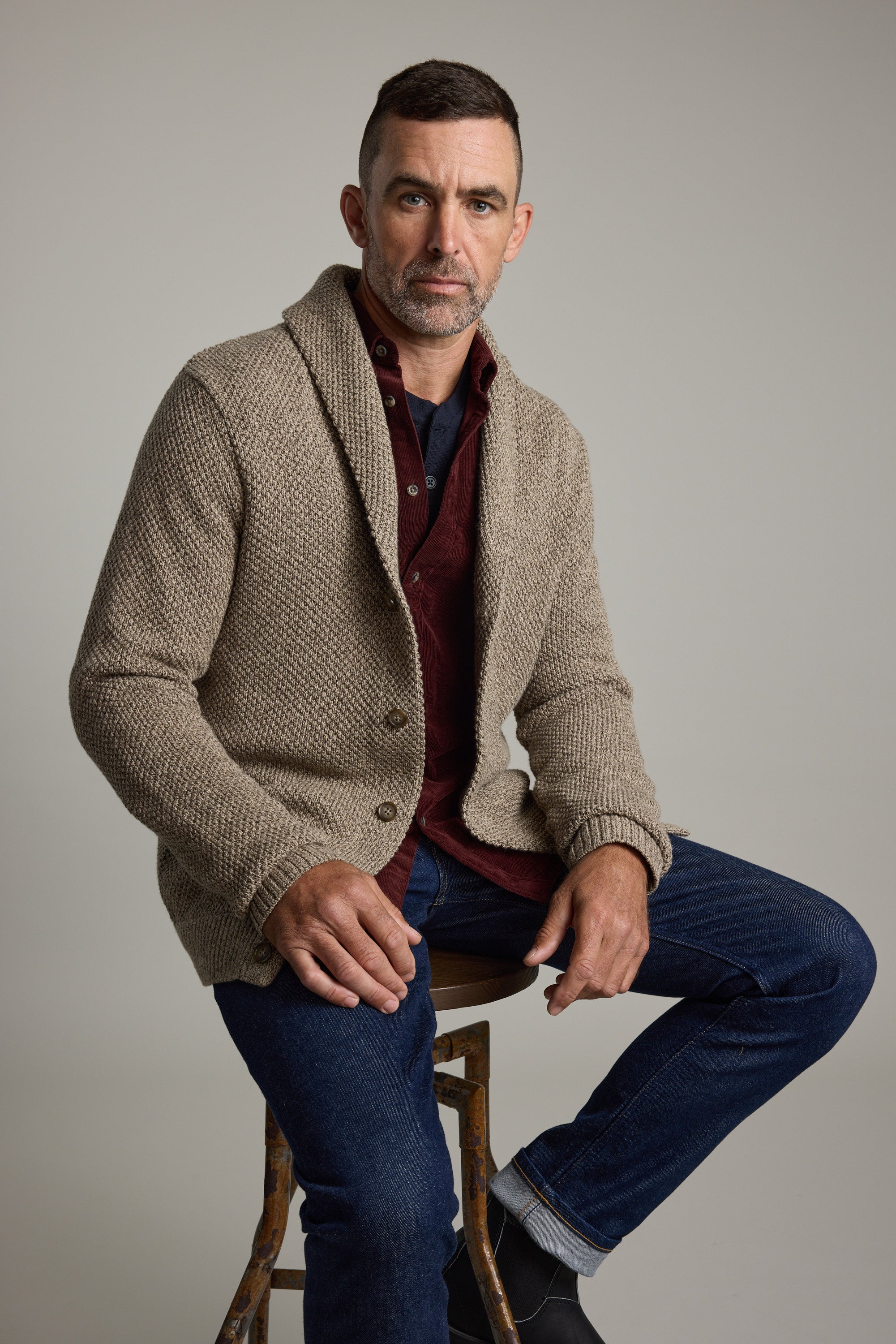 Man in Barkers Rudder Cotton Shawl Cardi and jeans sits on a stool, ready for winter layering.