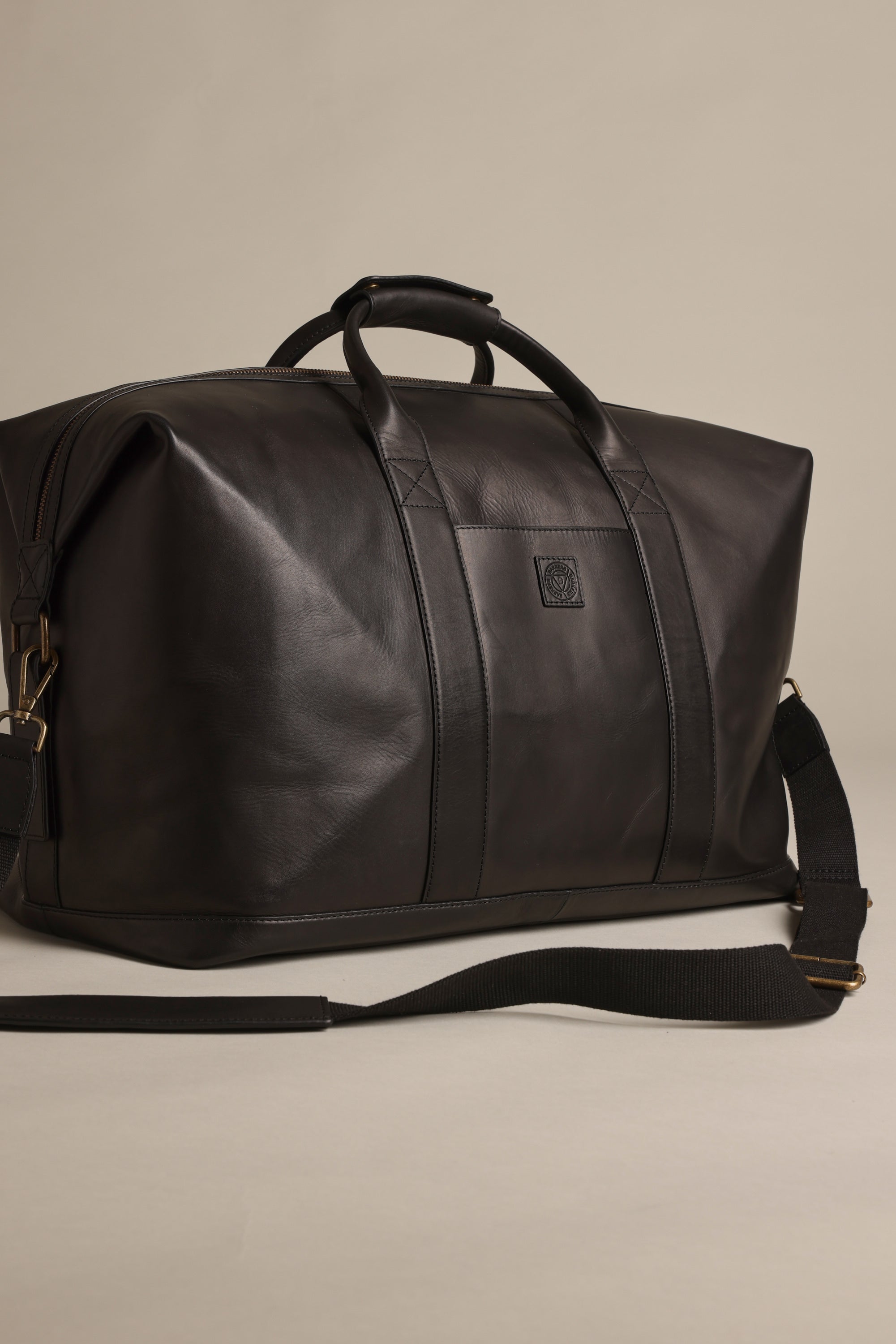 The Barkers Costello Leather Weekender is a large black premium leather bag featuring short handles, metallic hardware, and a detachable shoulder strap, shown on a plain beige background.