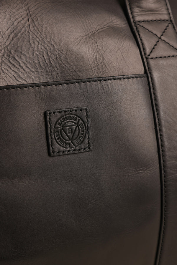 Close-up of the Barkers Costello Leather Weekender in black, showing premium grain, visible stitching, and a square embossed logo with circular text and shield—highlighting its quality craftsmanship.