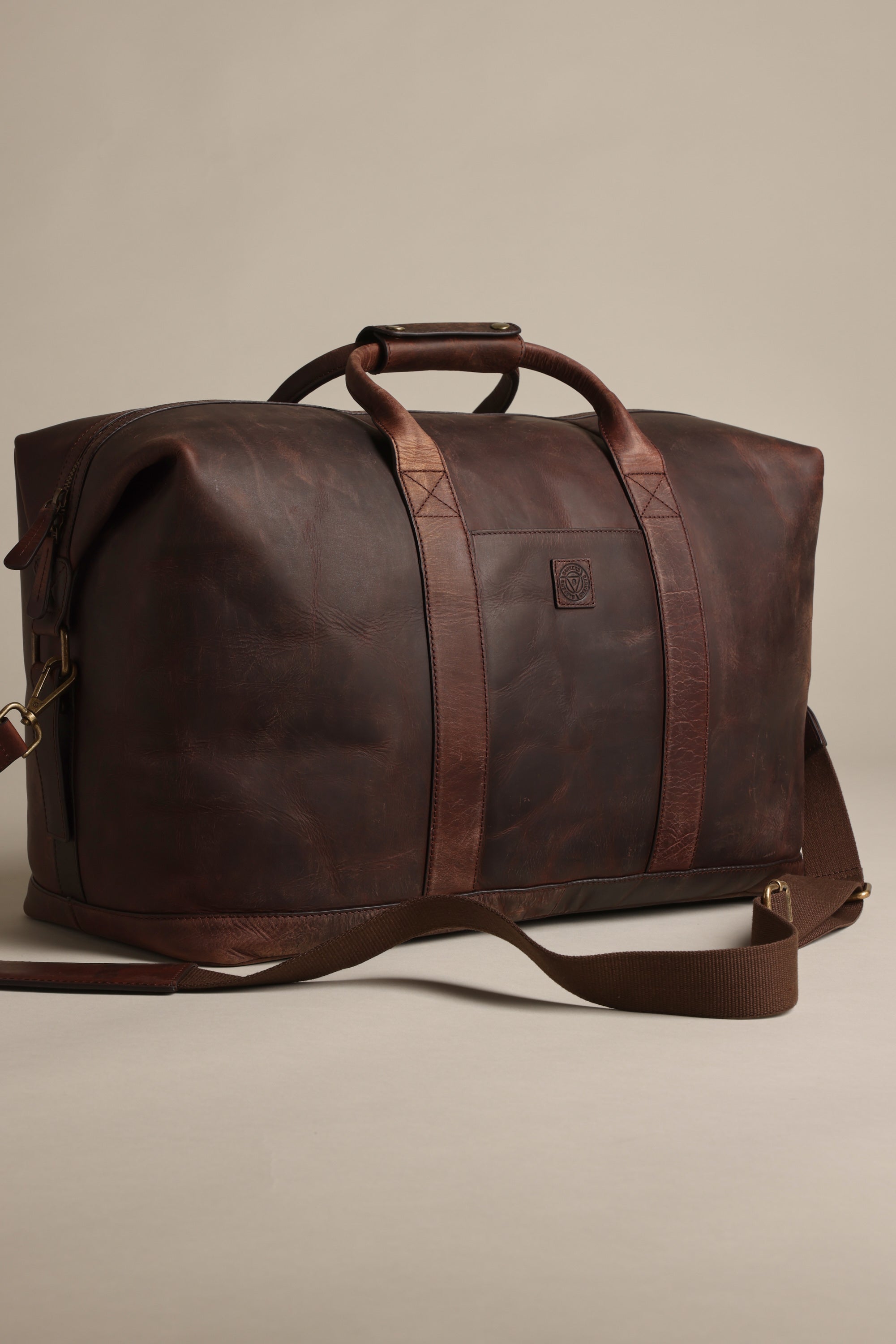 The Costello Leather Weekender by Barkers is a large, dark brown bag with two handles, a detachable shoulder strap, and brass hardware—ideal premium leather luggage for stylish getaways, shown against a plain beige background.
