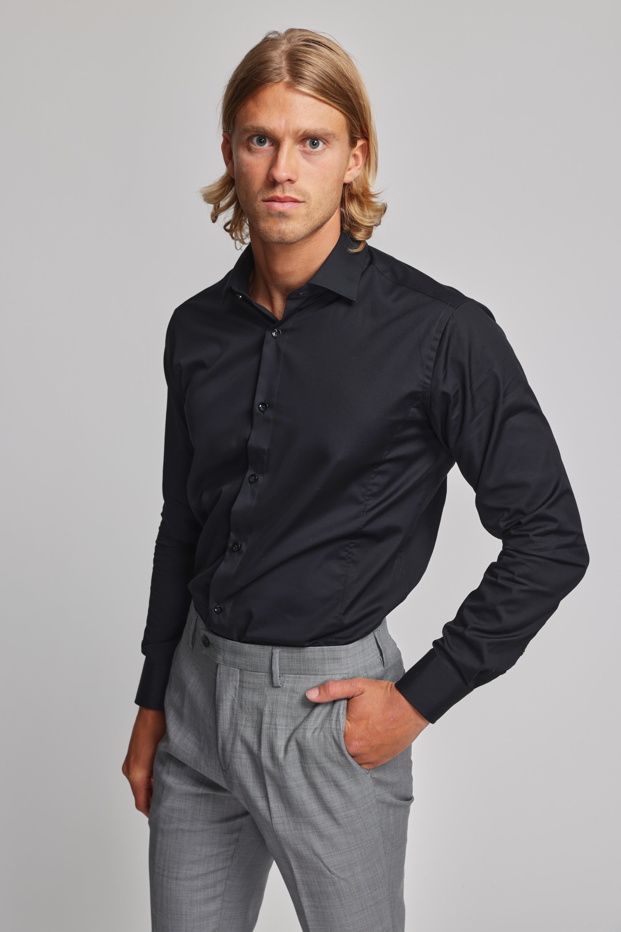 A man with shoulder-length blonde hair models the Barkers Tribeca Reg Cuff Shirt in black, tucked into light gray trousers. He stands against a plain backdrop, one hand in his pocket, gazing neutrally at the camera.