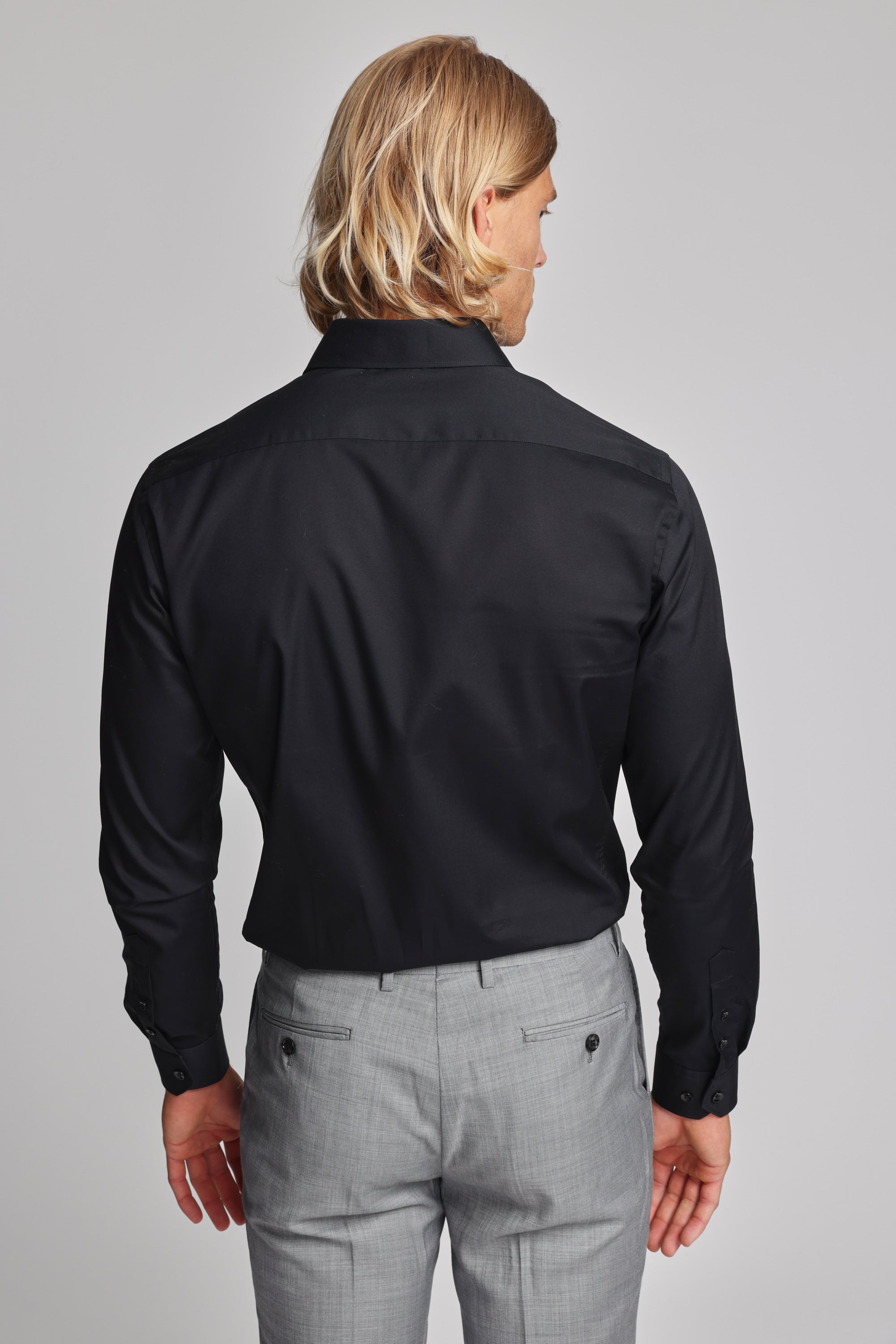 A person with shoulder-length blond hair wears a Barkers Tribeca Reg Cuff Shirt in black and light gray tailored dress pants, standing facing away from the camera against a plain light background.