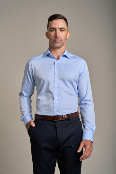 A man wearing the Barkers Tribeca Reg Cuff Shirt stands against a plain background with one hand in his pocket, paired with dark trousers and a brown belt, and maintains a neutral expression.