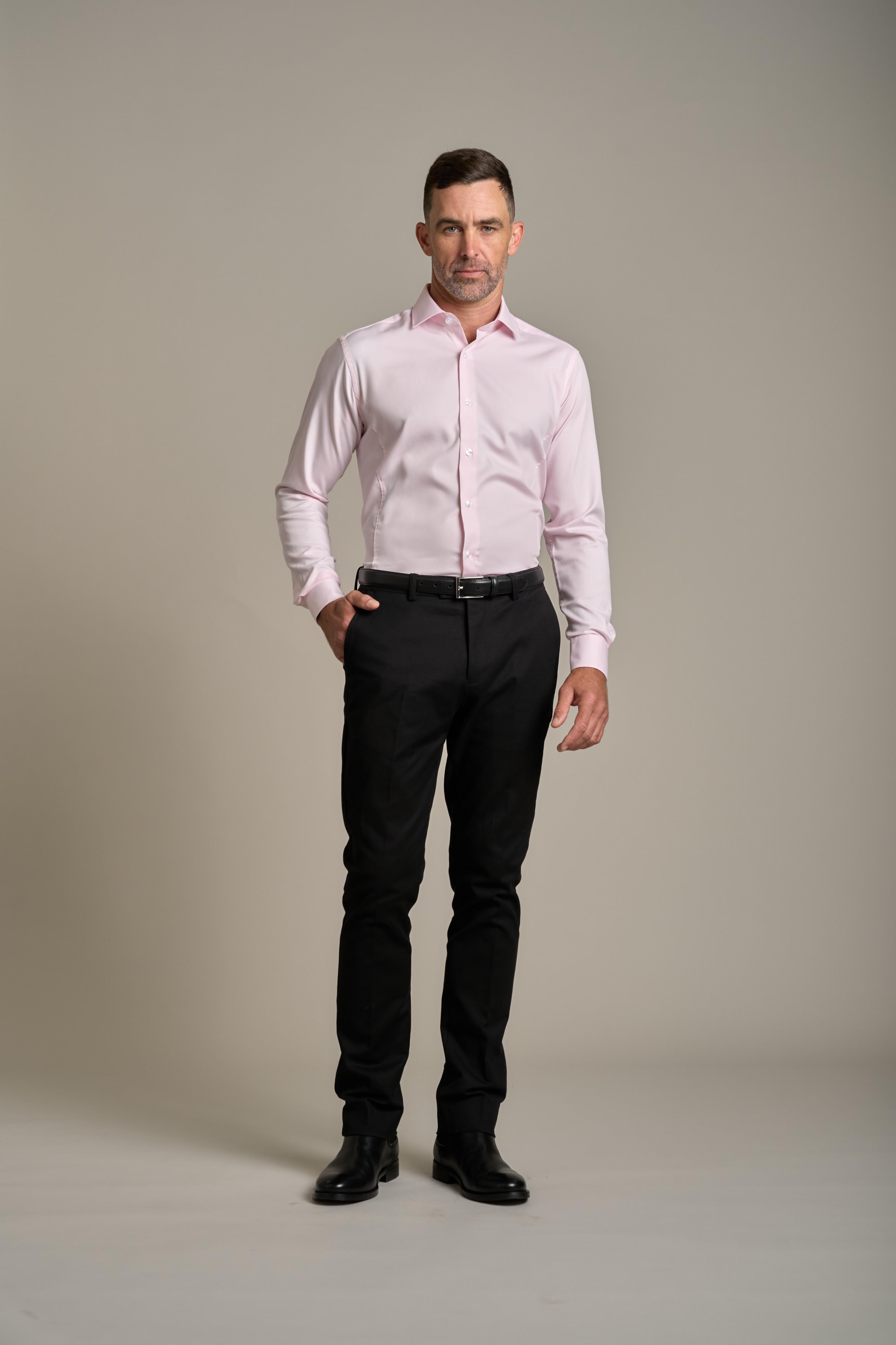 A man stands against a plain light background, wearing the Tribeca Reg Cuff Shirt by Barkers in light pink. He pairs it with black pants and shoes, has short dark hair, a neutral expression, and one hand in his pocket.