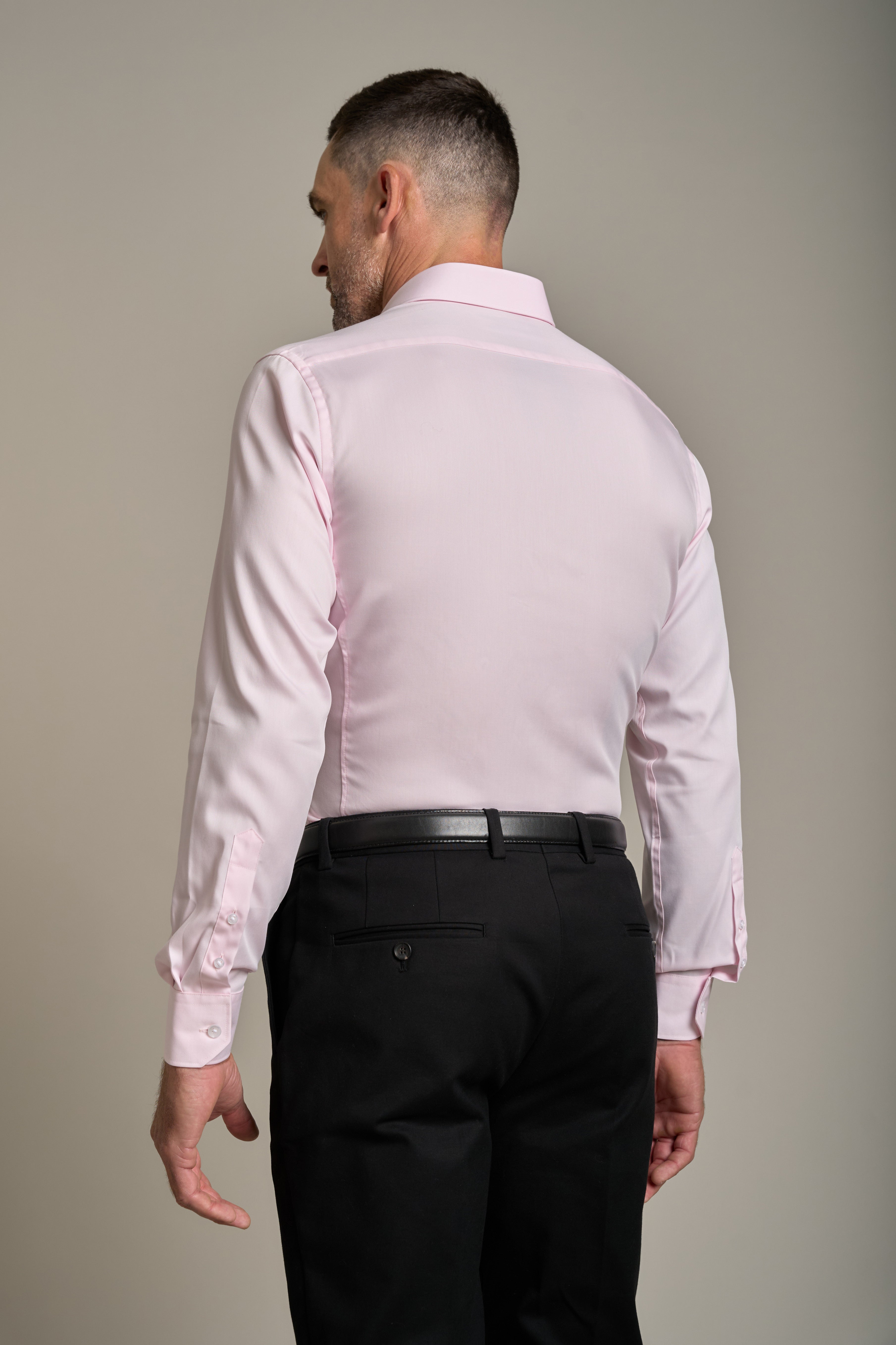 A man faces away from the camera, wearing a light pink Barkers Tribeca Reg Cuff Shirt in a tailored fit, tucked into black dress pants with a black belt. The background is plain and neutral.