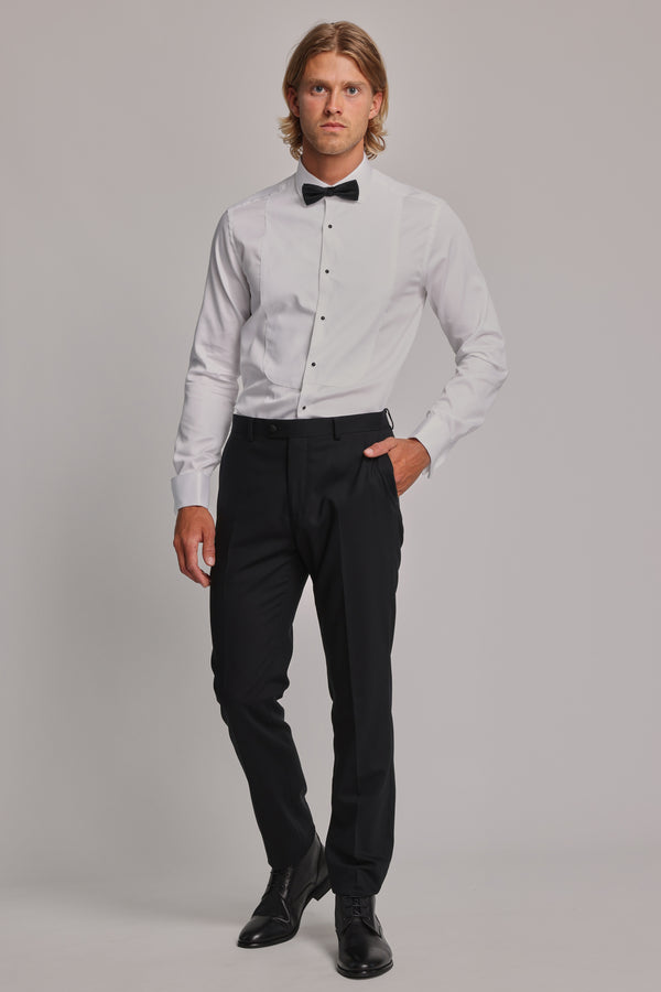 A man wears the Barkers Eldridge Dress Shirt—a white formal shirt with a tuxedo bib—paired with a black bow tie, dress pants, and shoes. He stands against a plain background, one hand in his pocket and wearing a neutral expression.