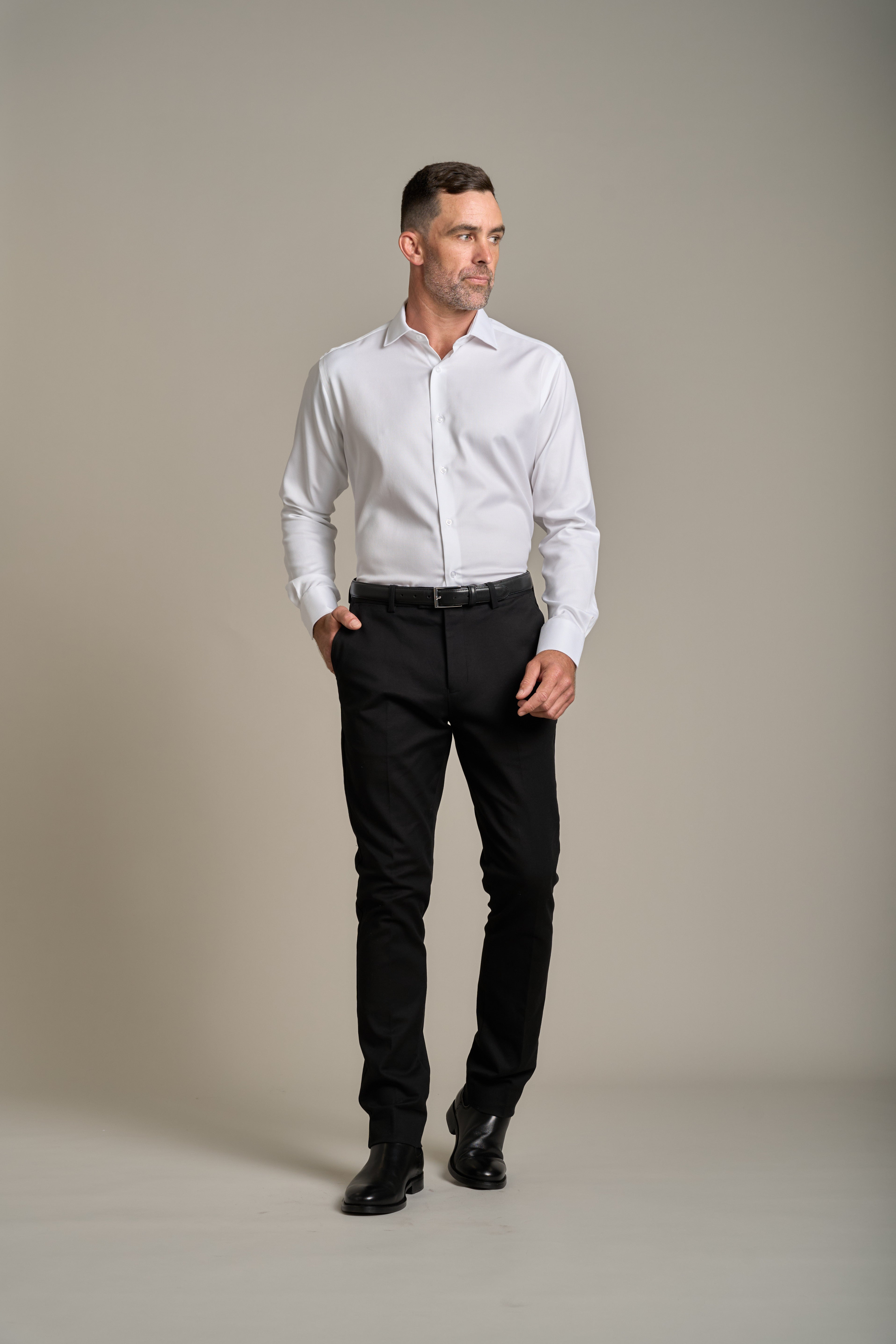 A man wearing the Barkers Tribeca Reg Cuff Shirt, black trousers, and black shoes stands against a plain light-grey background, looking to his left with one hand in his pocket.