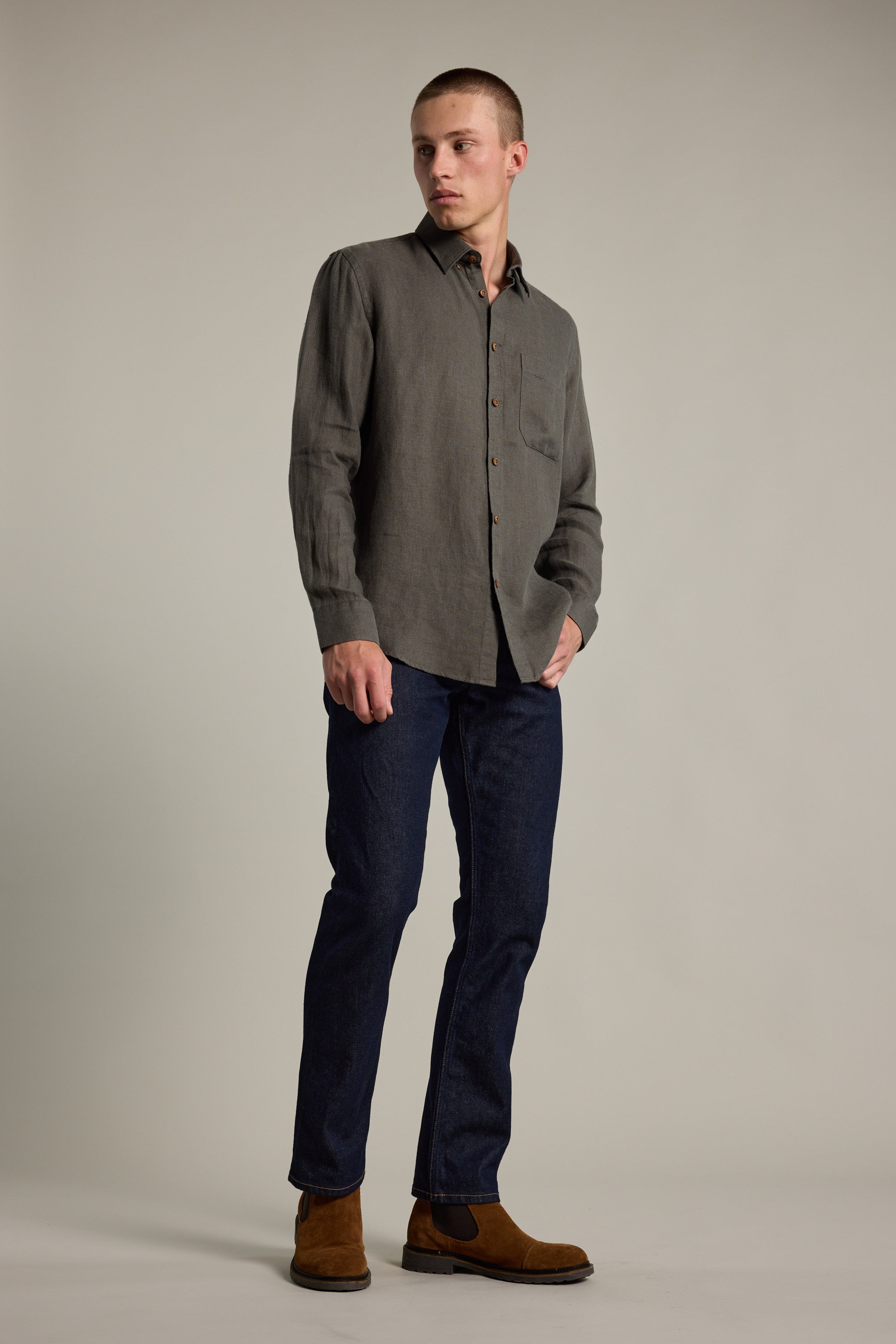 A person stands in a neutral studio wearing the Barkers Delano Linen Shirt in gray, paired with dark blue jeans and brown suede boots. They have short hair, look to the side, and keep one hand in their pocket.