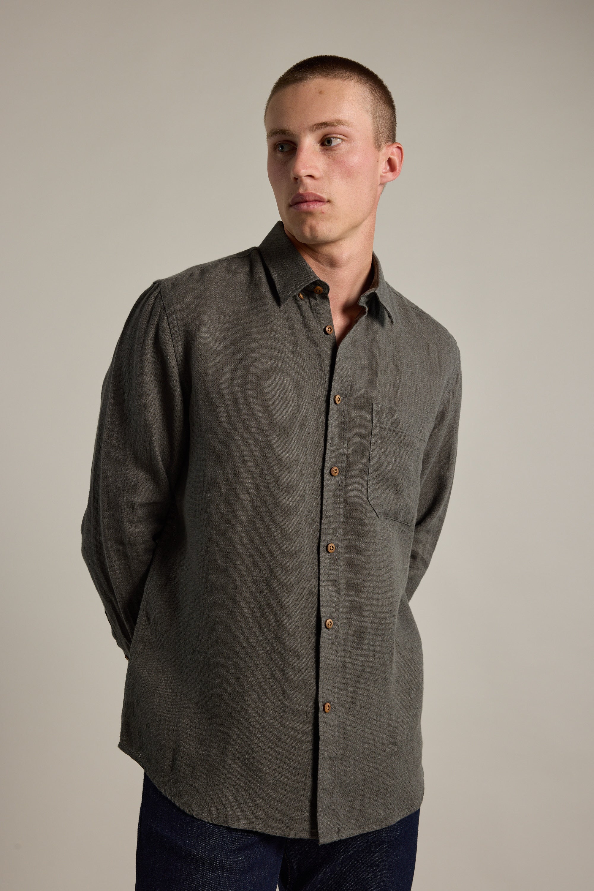 A young man with short hair wears the Delano Linen Shirt by Barkers—a classic fit, long-sleeved dark gray shirt with a chest pocket—standing against a plain light background and looking slightly to his left.