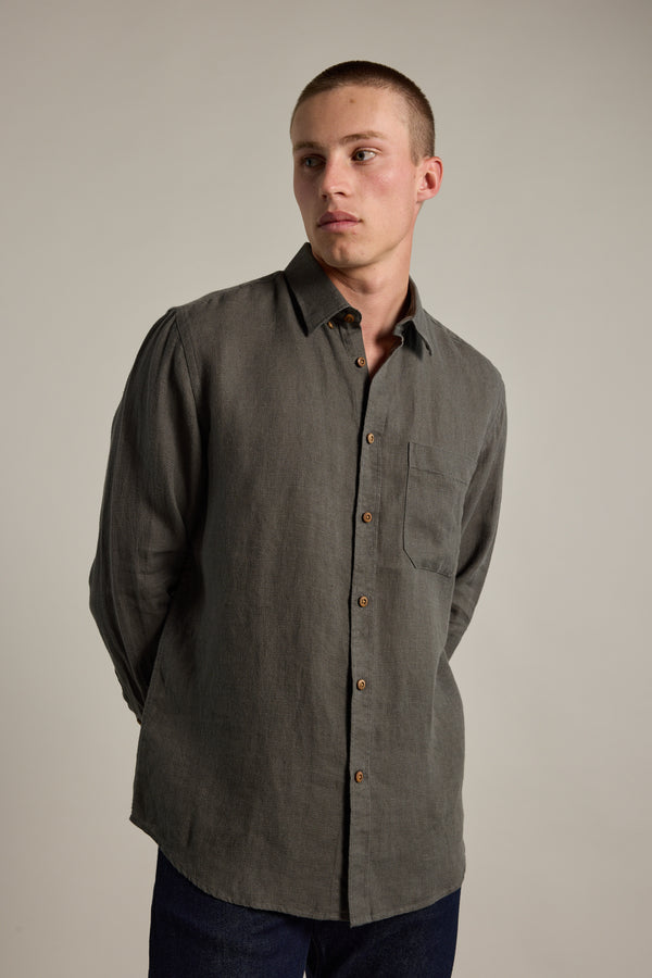 A young man with short hair wears the Delano Linen Shirt by Barkers—a classic fit, long-sleeved dark gray shirt with a chest pocket—standing against a plain light background and looking slightly to his left.