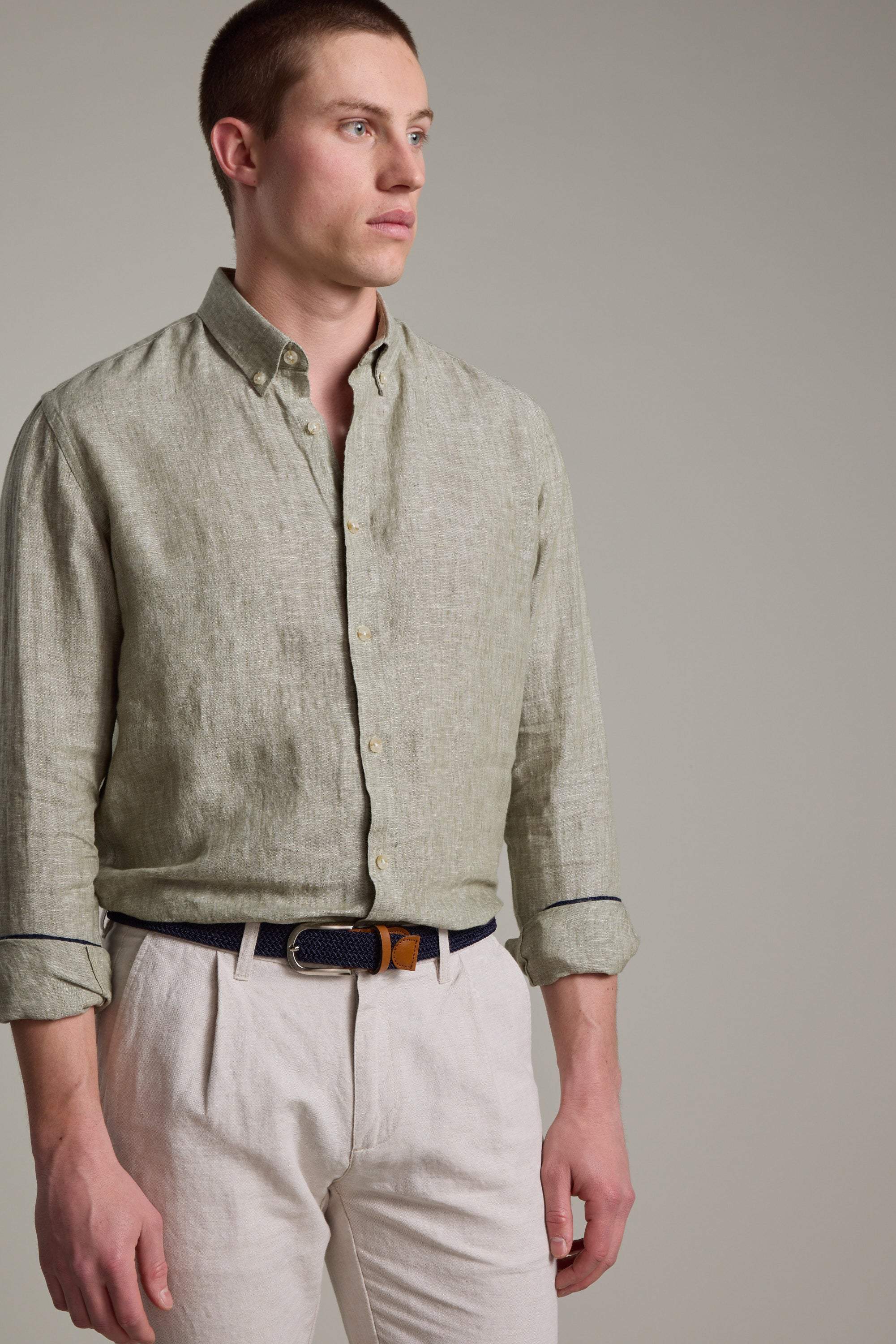 A man wears the Barkers Dorset Linen Shirt in light green—breathable French linen with a tailored fit, sleeves rolled up—paired with beige trousers, standing against a plain light gray background and looking to the side.