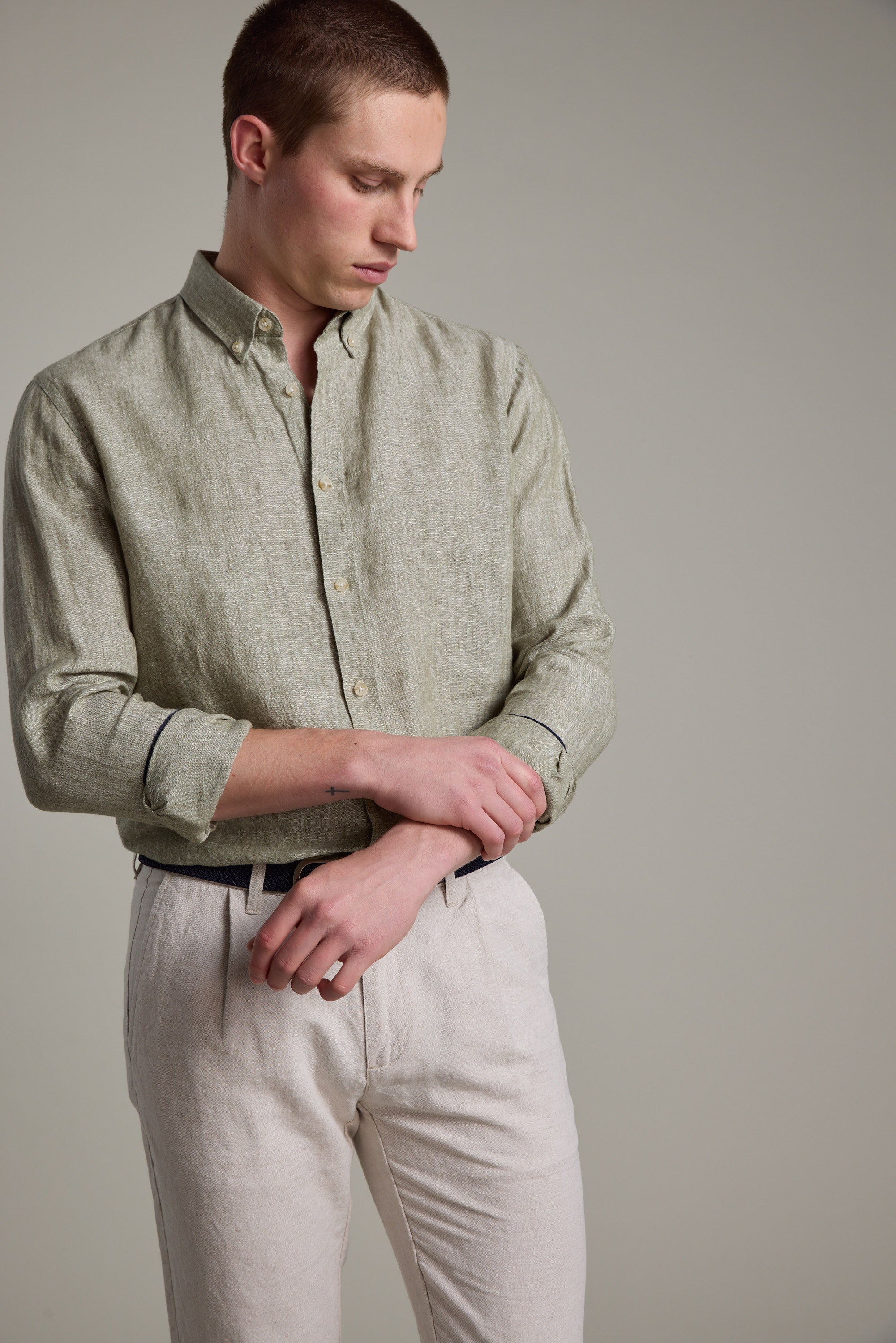 A young man in a light green Barkers Dorset Linen Shirt with a tailored fit and light beige pants stands against a plain background, looking down and adjusting his shirt sleeve.
