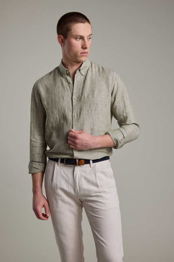 A young man with short hair wears the Barkers Dorset Linen Shirt in light olive-green, tailored fit, paired with beige trousers and a black belt. He stands against a plain light gray background, looking to his left with one hand by his side.