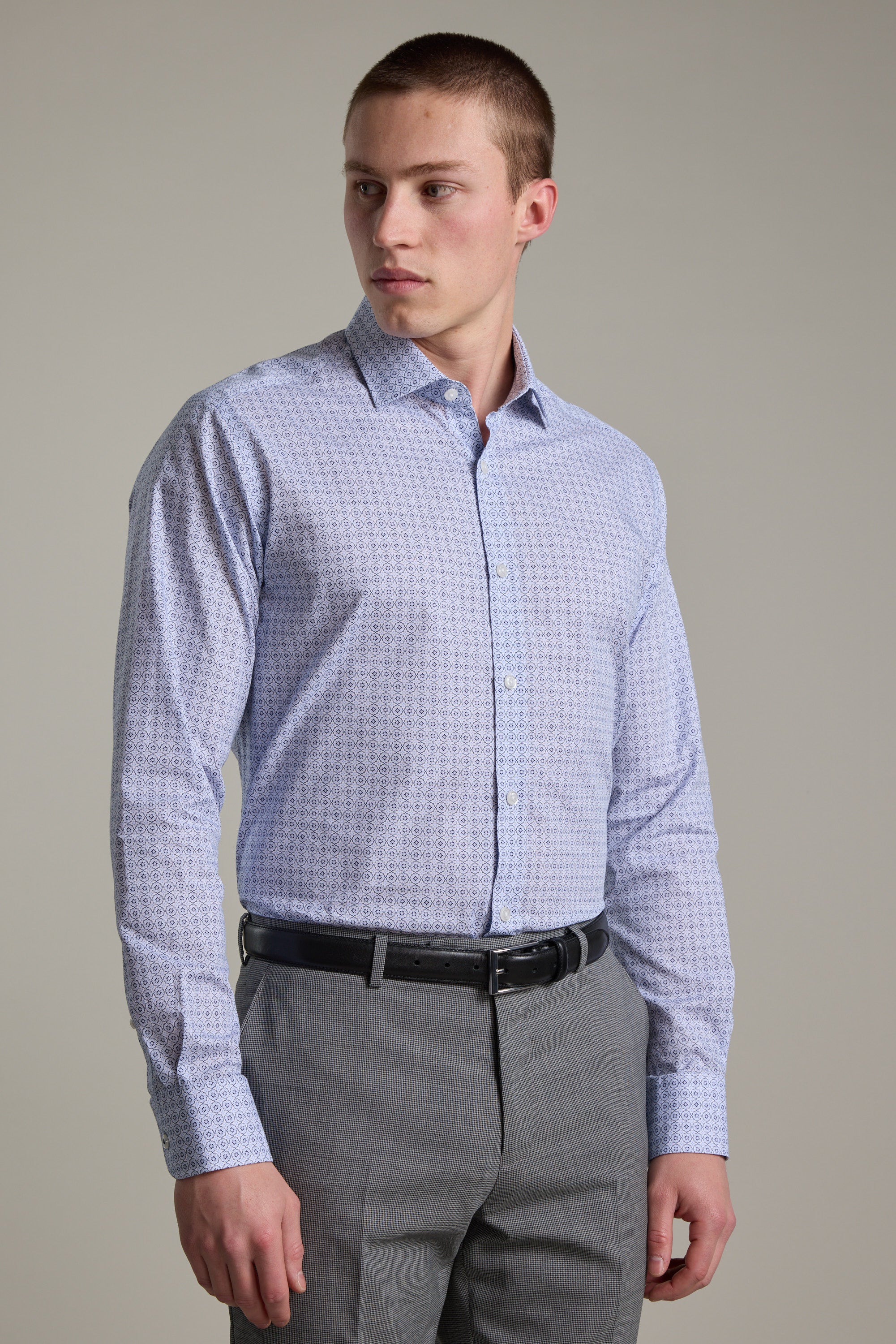 A man wearing the Barkers Seton Print Business Shirt in light blue stands against a plain background, paired with gray trousers and a black belt, looking to his left.