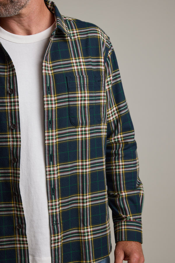 A person wears a white T-shirt under an open, green and yellow Barkers Brennan Check Overshirt. Only the lower half of the face, torso, and right arm are visible against a plain background. Made from responsibly sourced cotton.