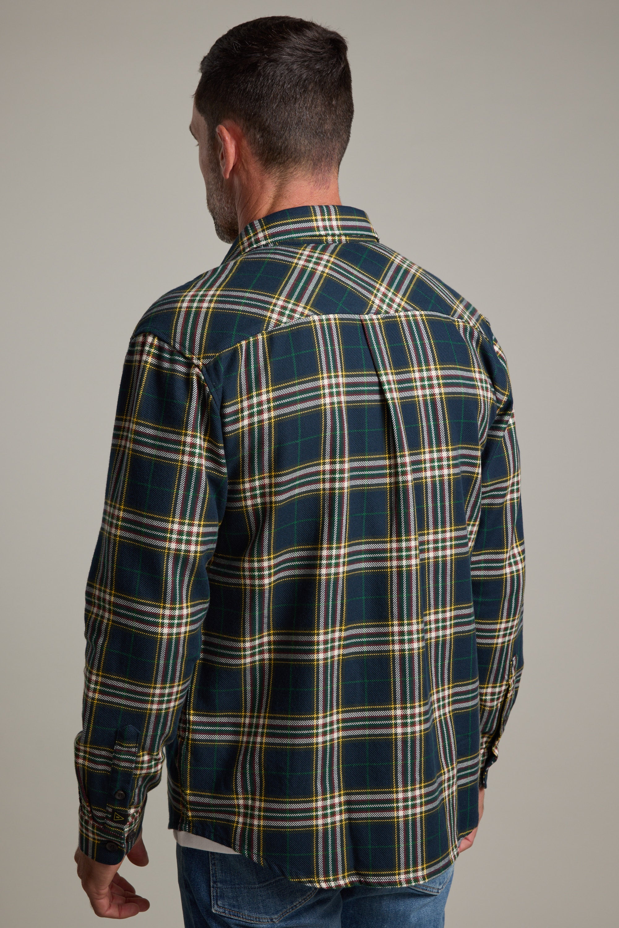 A man with short dark hair, seen from behind, wears a green, yellow, and white Barkers Brennan Check Overshirt made from responsibly sourced cotton and blue jeans against a plain light background.