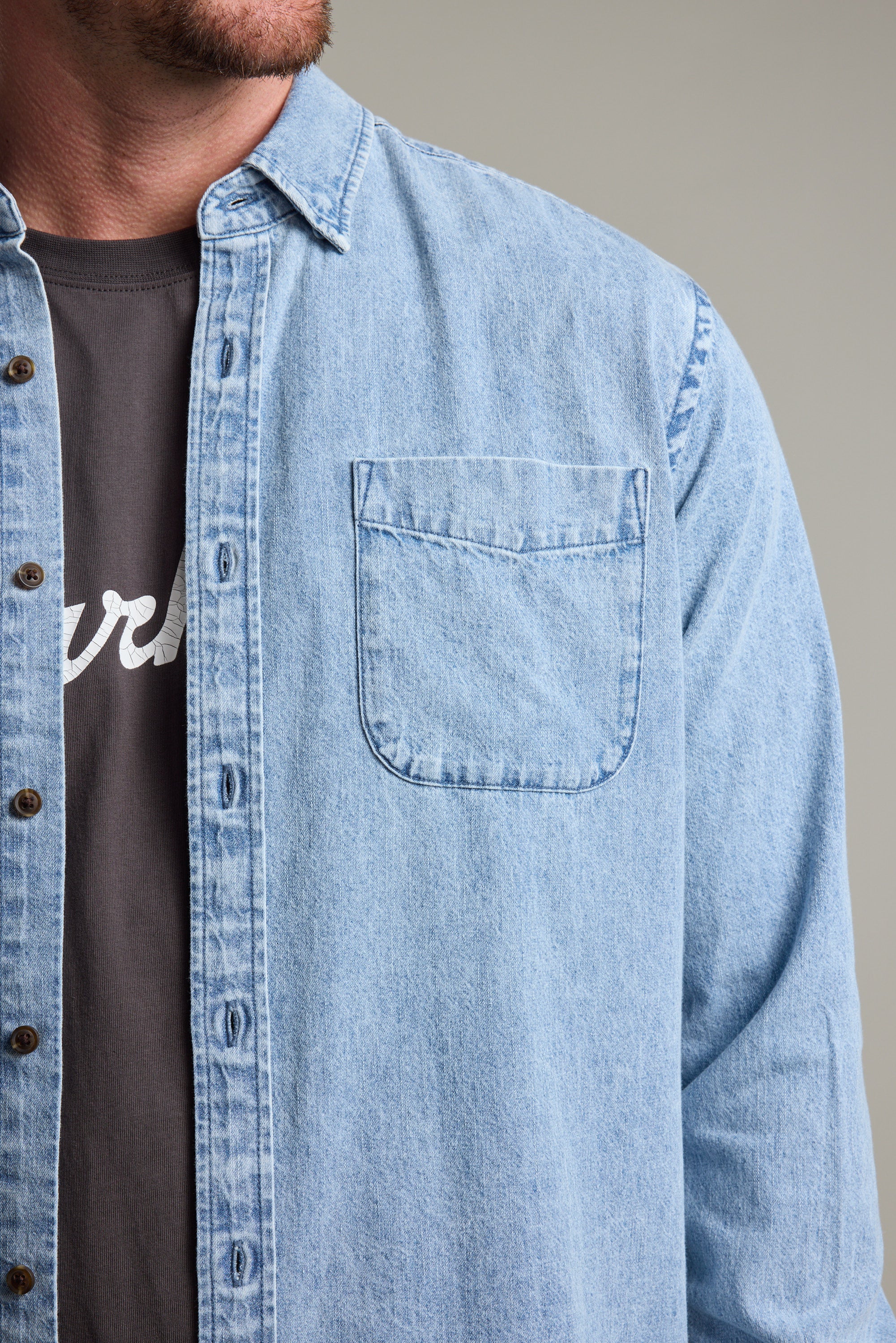 A man models the Barkers Ruxton Denim Shirt, a classic-fit light blue overshirt, layered over a dark tee with white lettering; only his face’s lower half, chest, and upper arm are visible.
