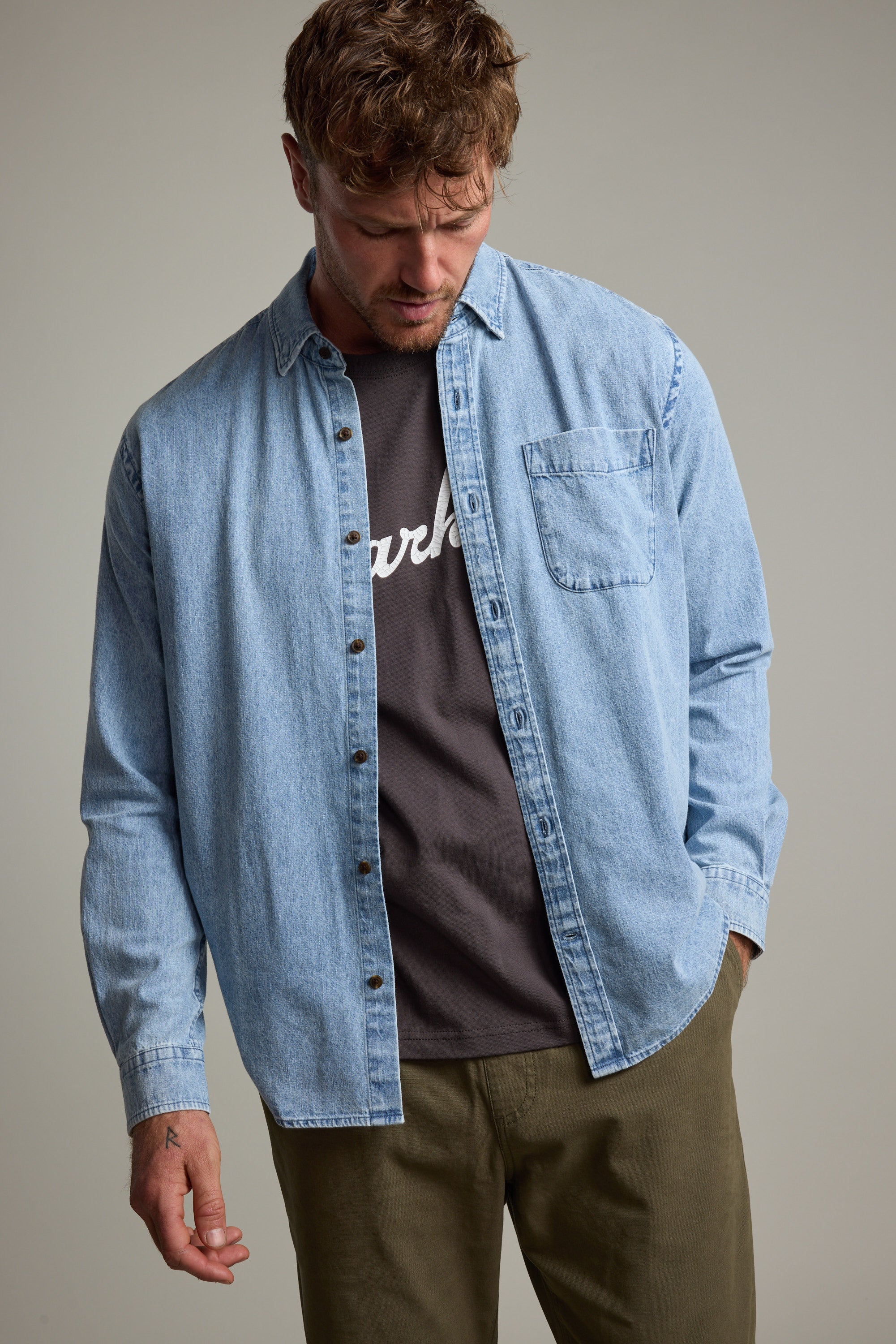 A man wears the Barkers Ruxton Denim Shirt in a classic fit, layered over a dark tee with white text and olive green pants, standing with head slightly bowed and one hand in his pocket against a neutral background.
