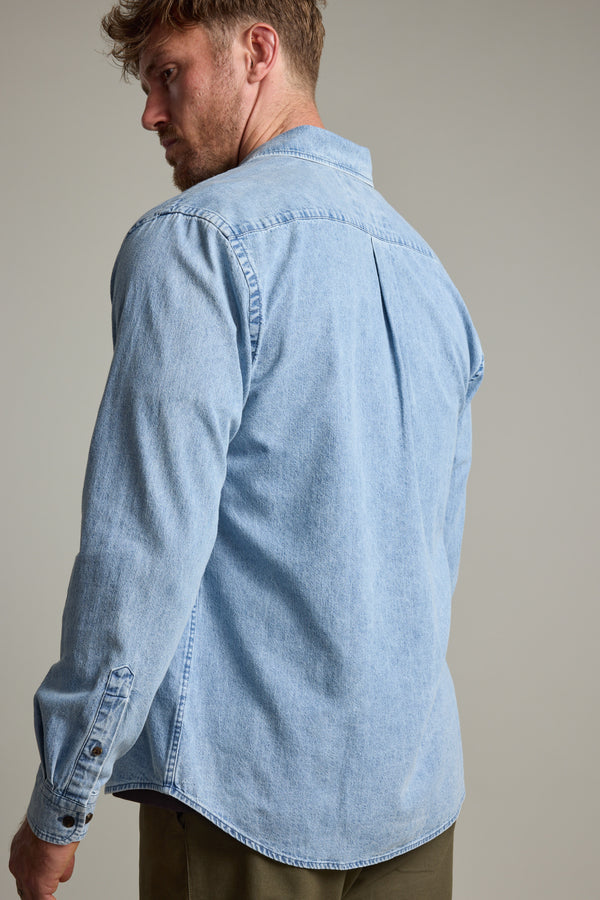 A man with light brown hair and a beard stands against a plain background, wearing the Barkers Ruxton Denim Shirt styled as an overshirt with olive green pants.