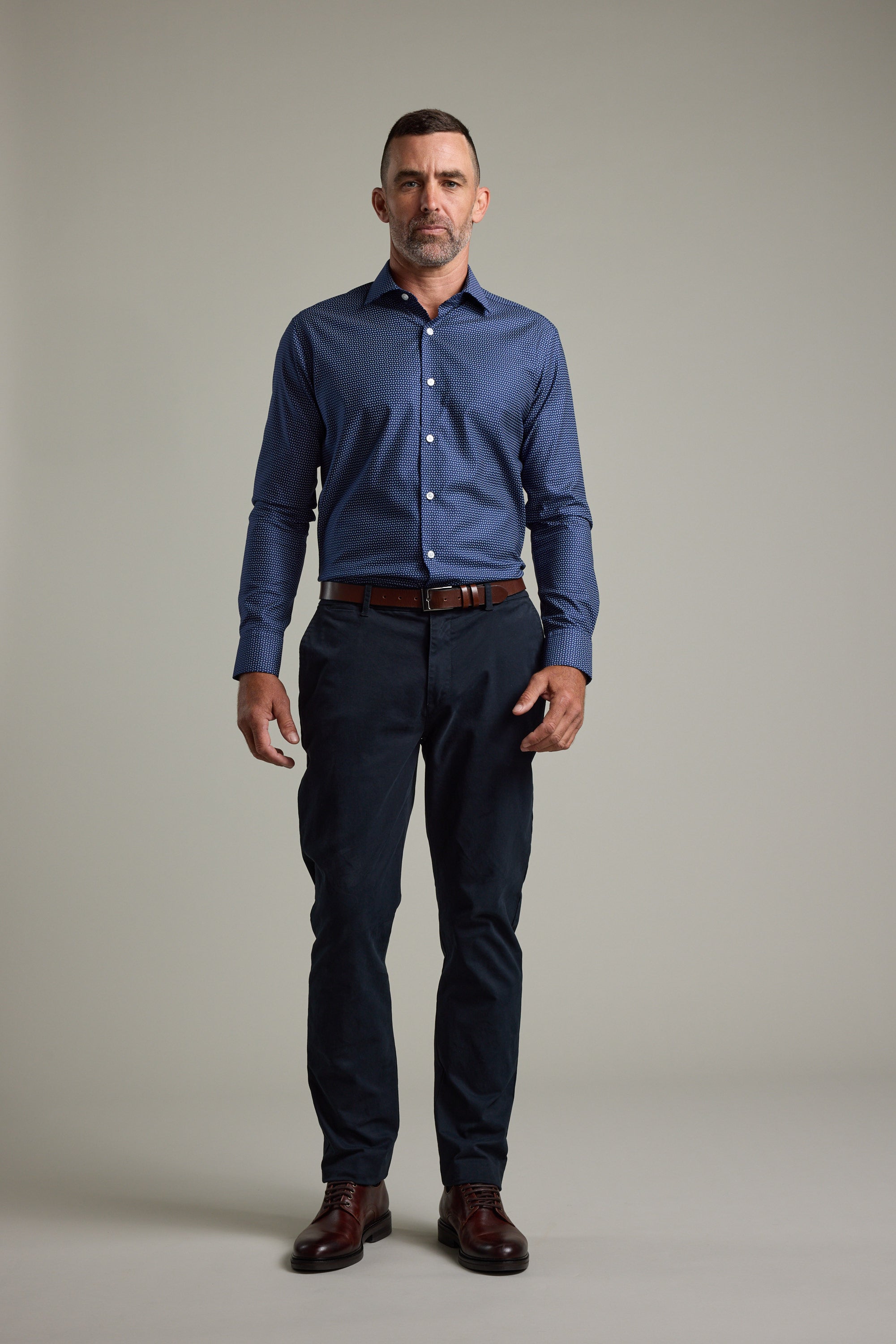A man with short hair and a trimmed beard stands against a plain background, wearing the Barkers Maiden Paisley Business Shirt, dark trousers, a brown belt, and brown shoes. He faces the camera with a neutral expression.