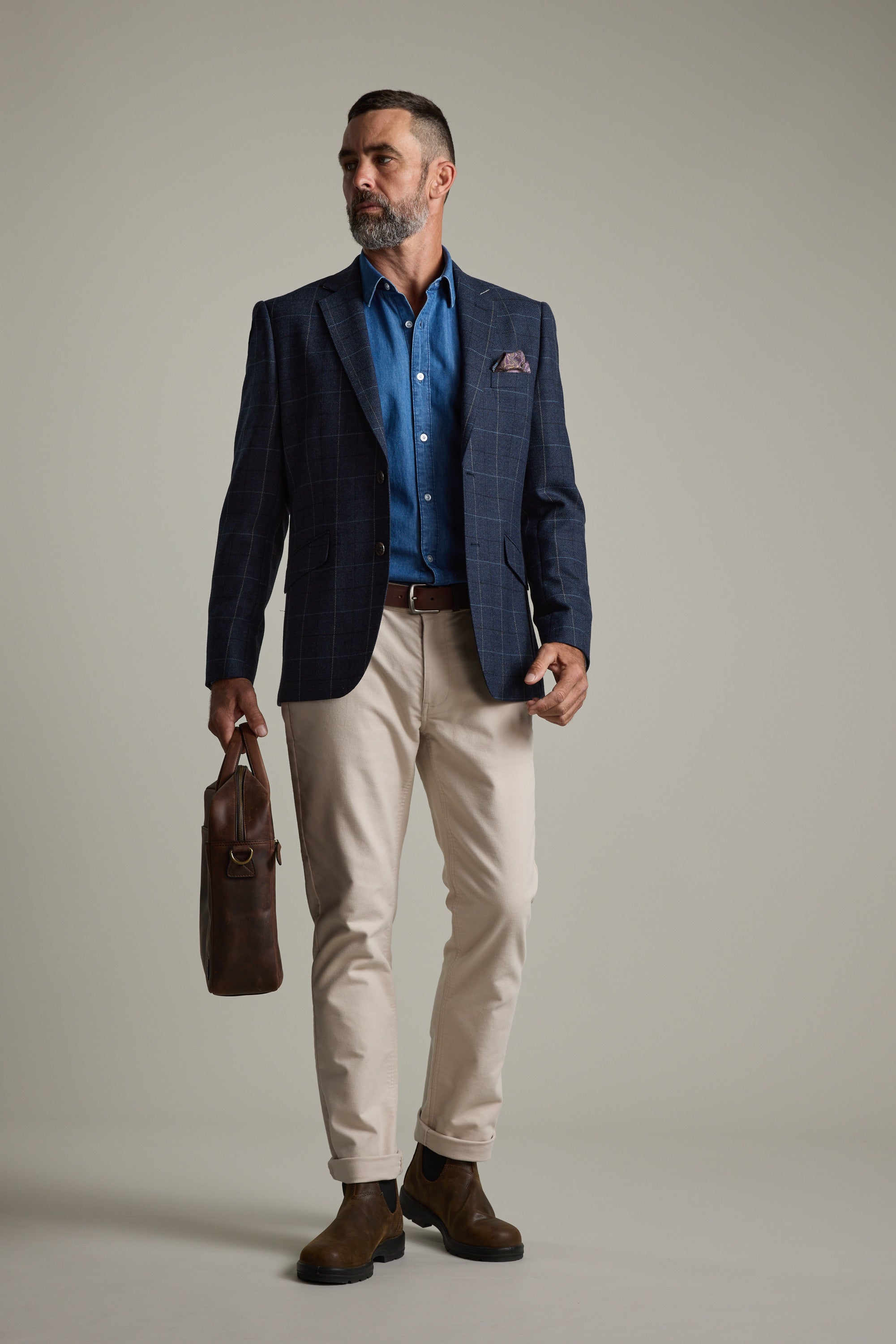 Man in Barkers Wyatt Denim Business Shirt, blazer, beige pants, holding brown bag on grey.